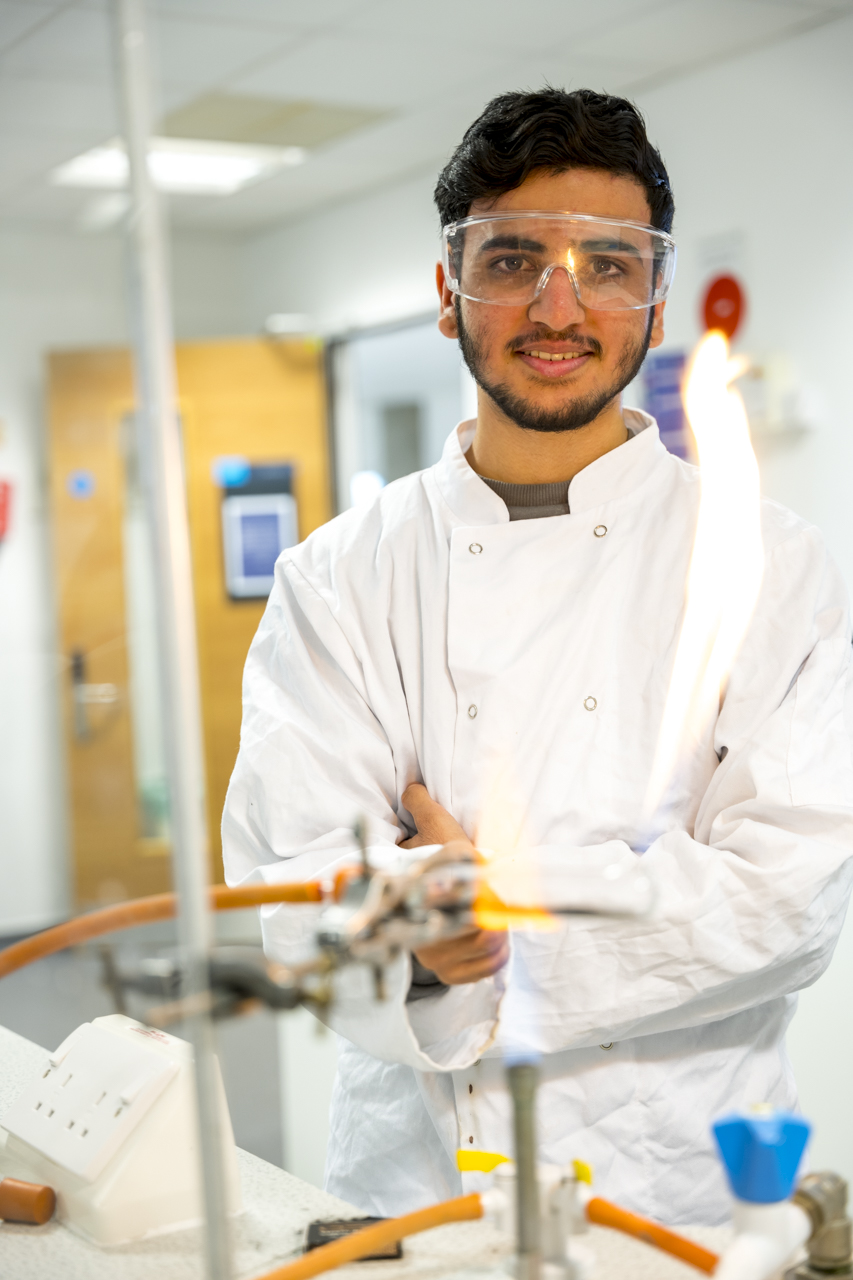 a science student wearing a white lab coat and protective glasses as he stands behind a bunsen burner