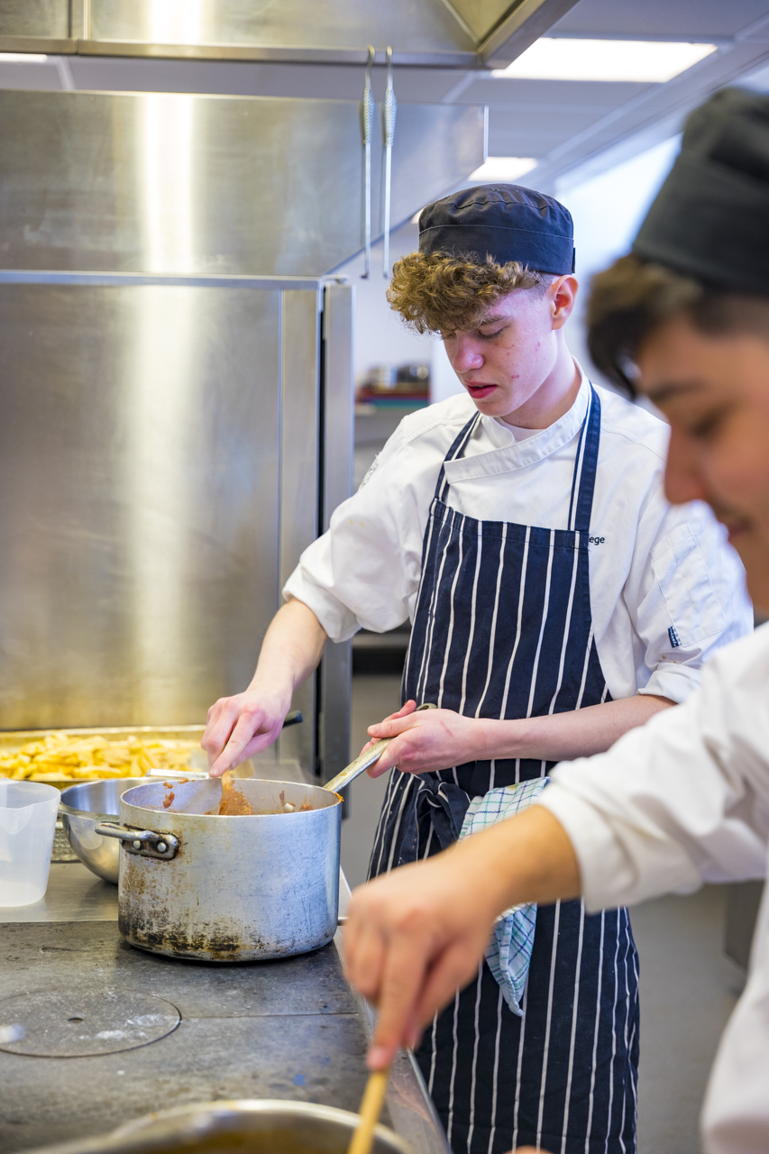 a catering student wearing an apron and hat working at a worktop stirring something in a pot