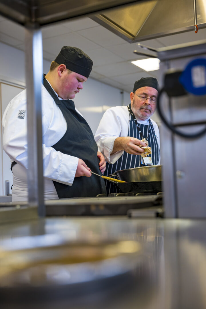 a catering tutor assisting a catering student in the kitchen as they prepare meals for guests