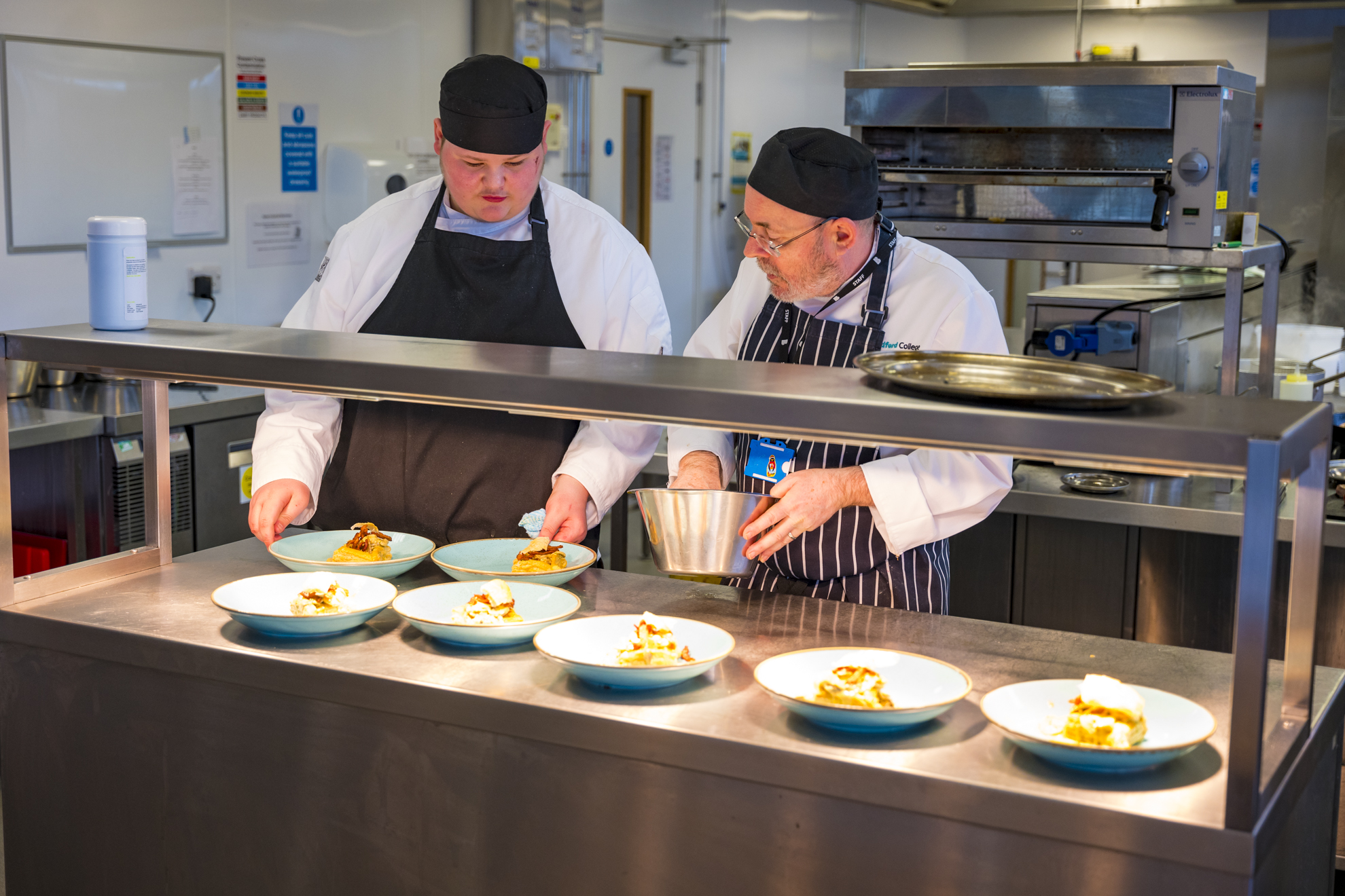 a catering tutor assisting a catering student in the kitchen as they prepare meals for guests