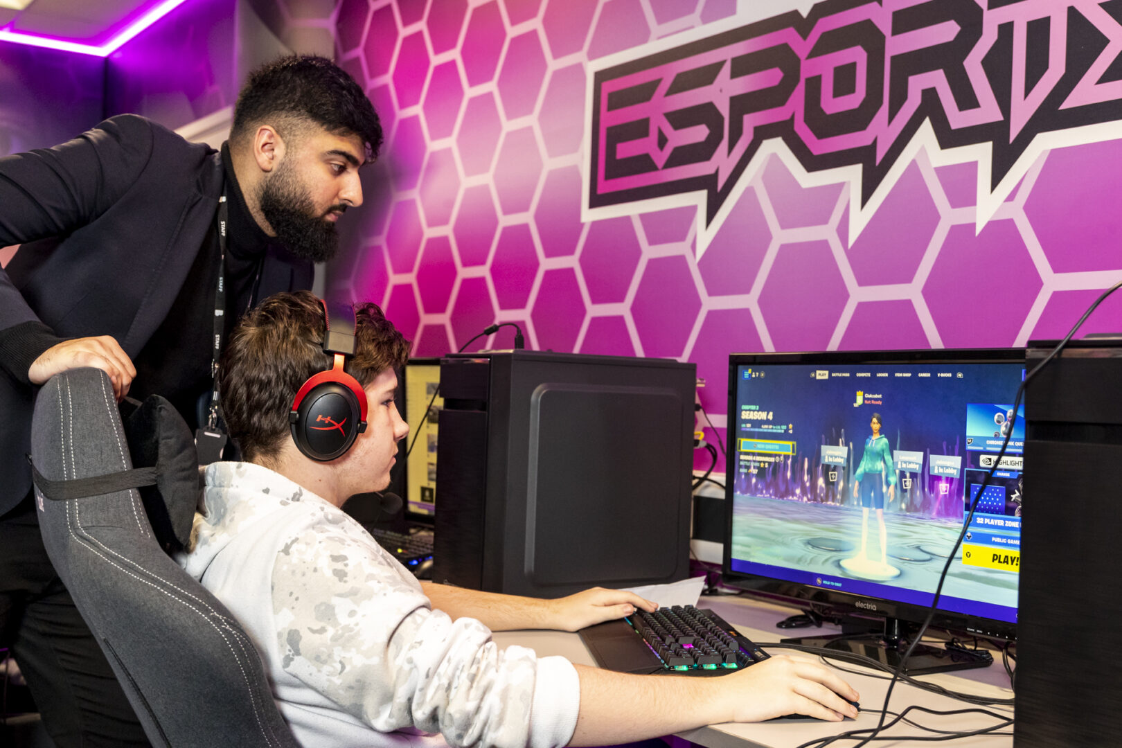 an esports student sits at a computer screen as a tutor looks over at his work