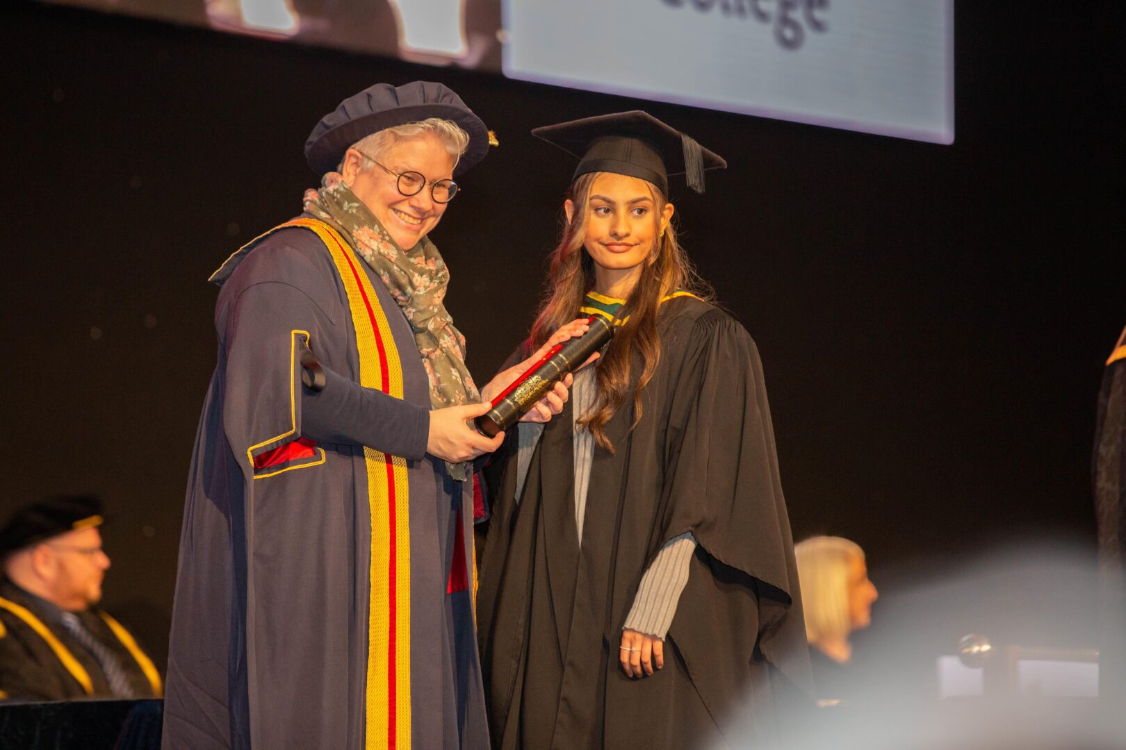 a smiling graduate collects their certificate on stage as they pose for a photograph