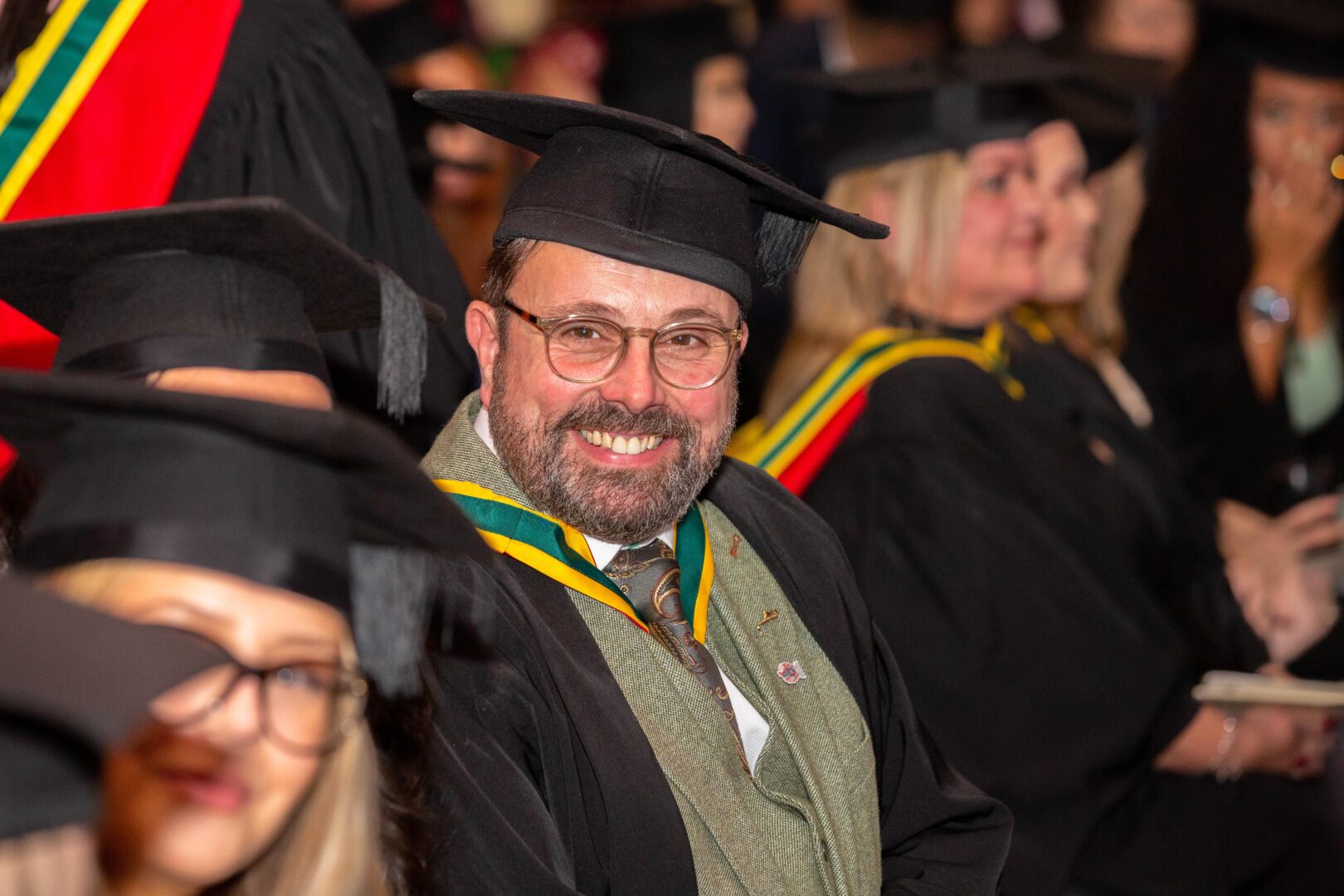 a smiling student sits among his peers and he waits to graduate