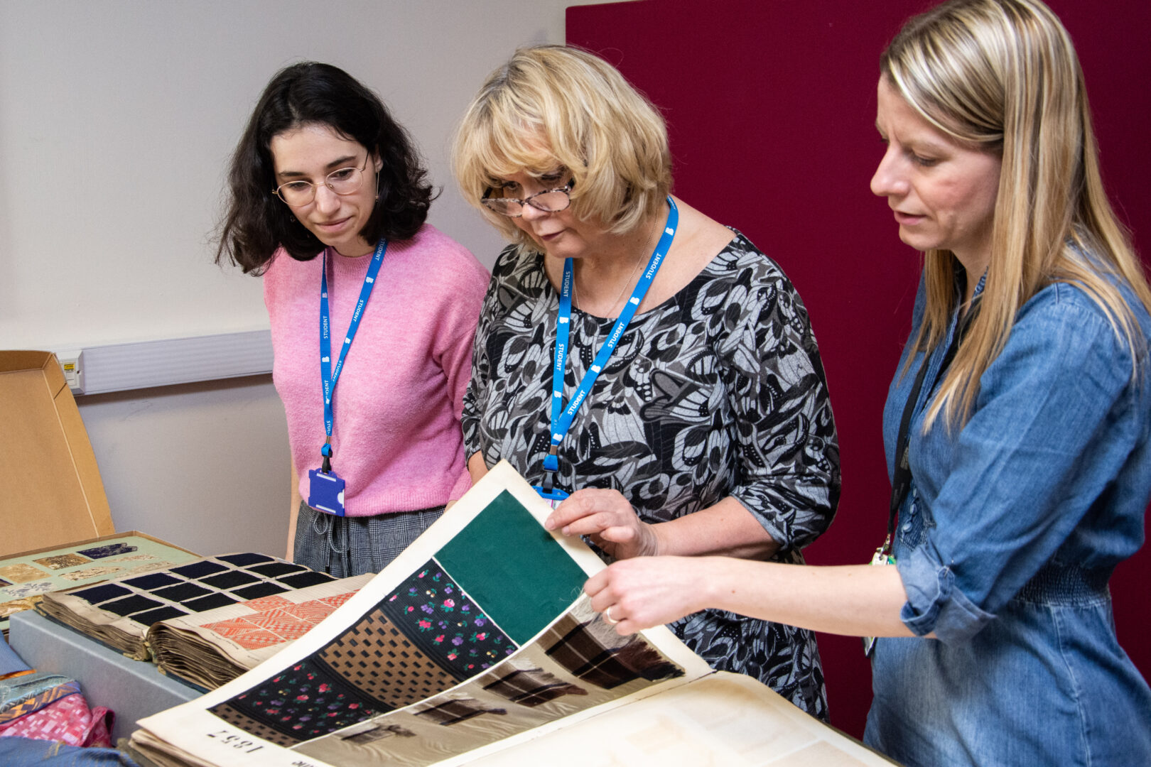 three textiles students visiting the bradford textiles archive, they are stood looking through a document filled with fabrics and textiles