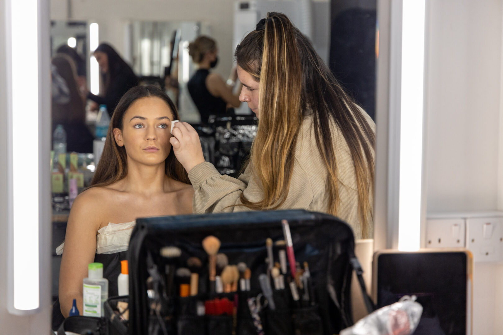 a model sits in front of a mirror as a student applies make-up to their face