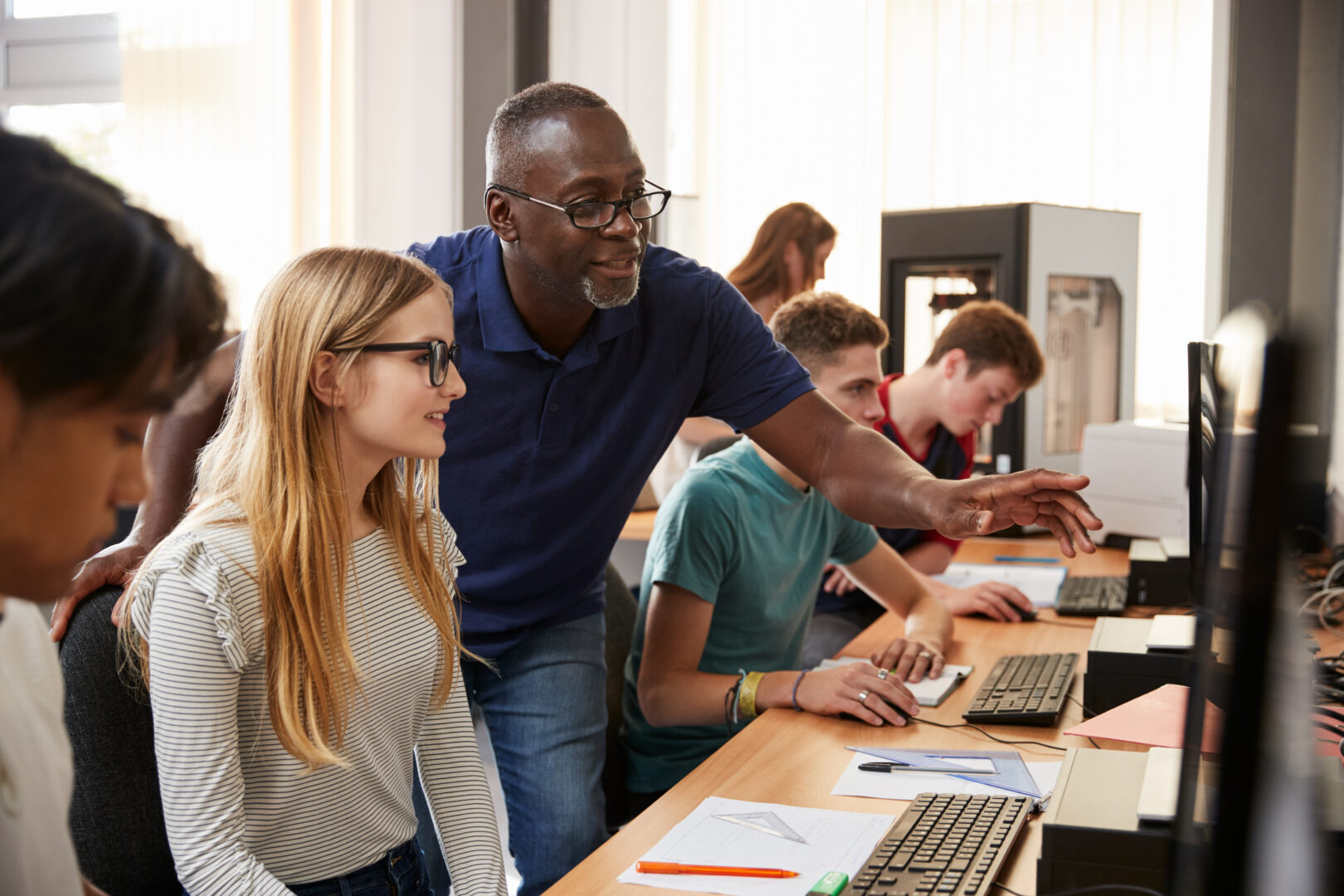 a teacher stands at a students desk as they look at a computer screen
