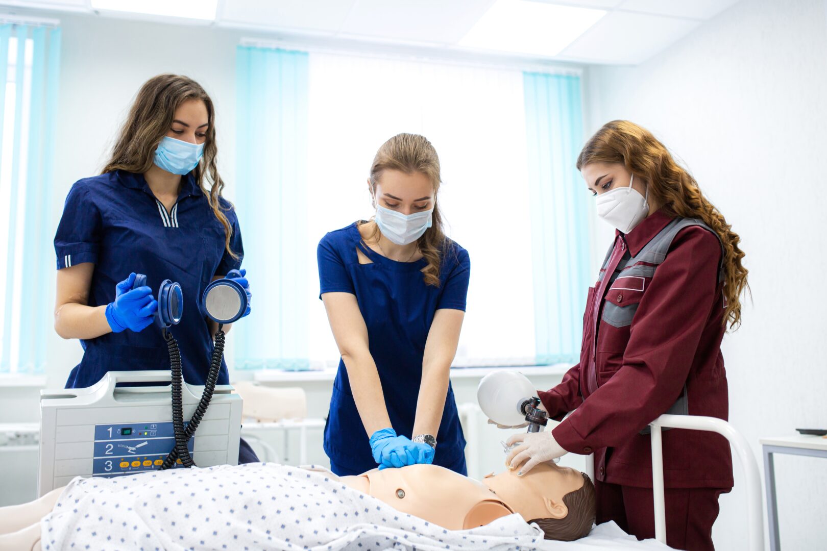 three healthcare students stand around a dummy patient, one student wearing a mask and gloves performs cpr on the doll
