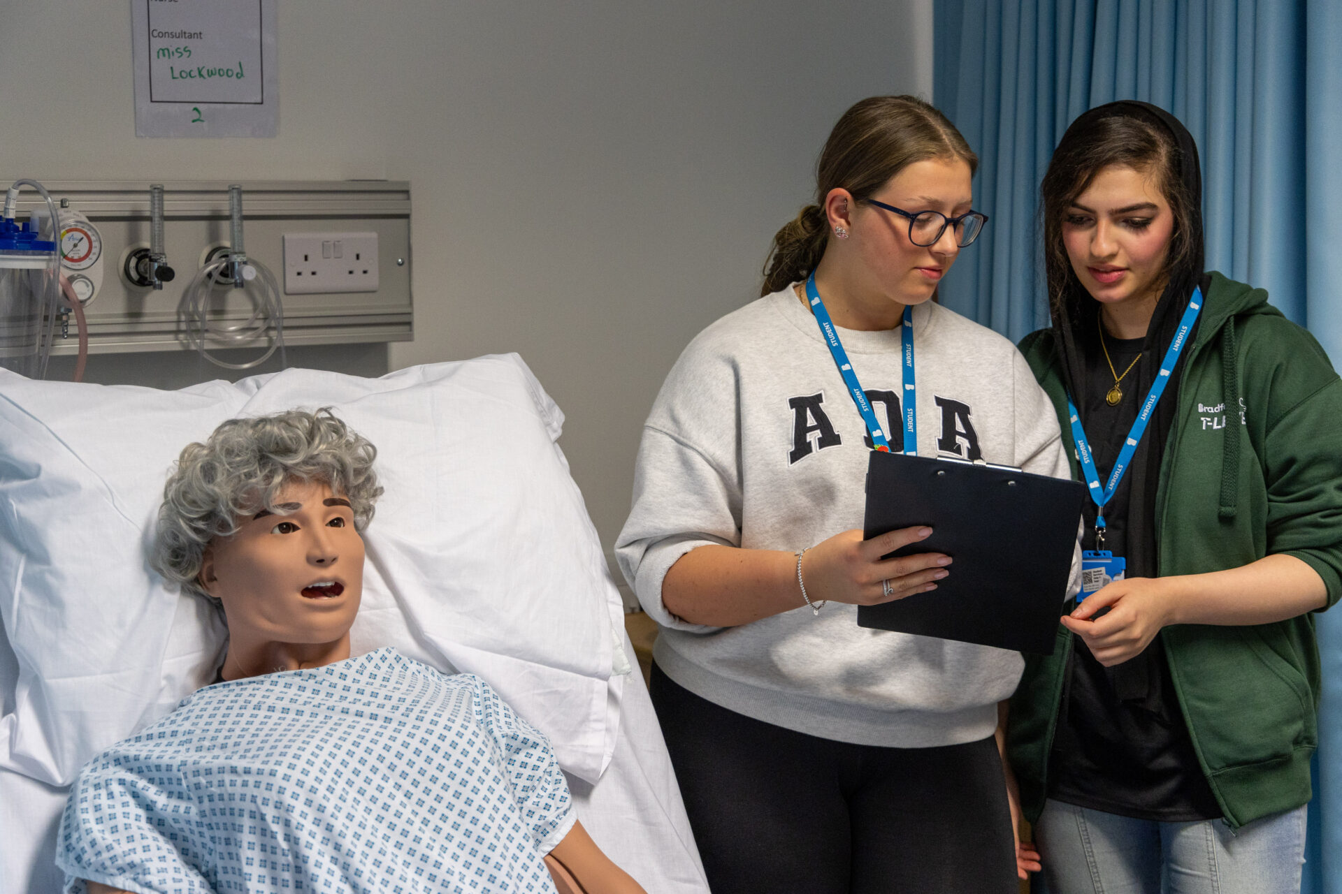 Two female students with clipboard stood beside training doll in our mock clinical ward