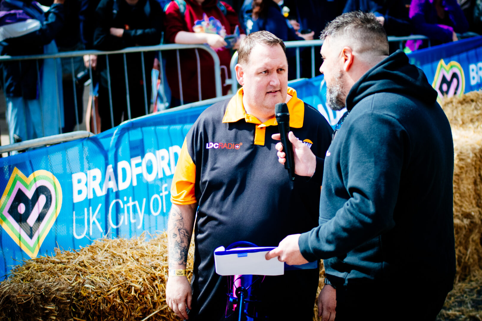 two men stand speaking, one is holding a microphone