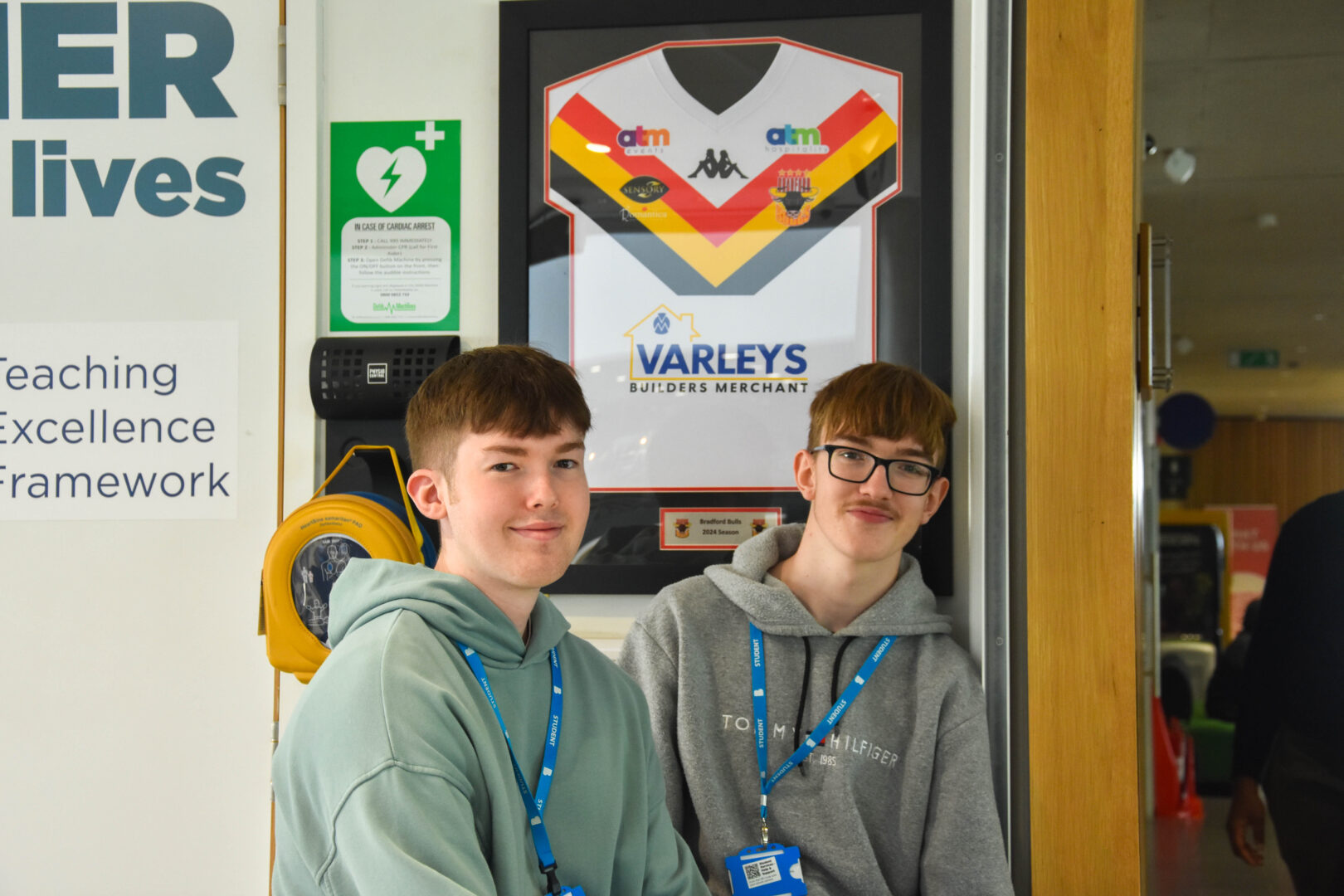 two bradford college students stand smiling next to a framed bradford bulls rugby shirt