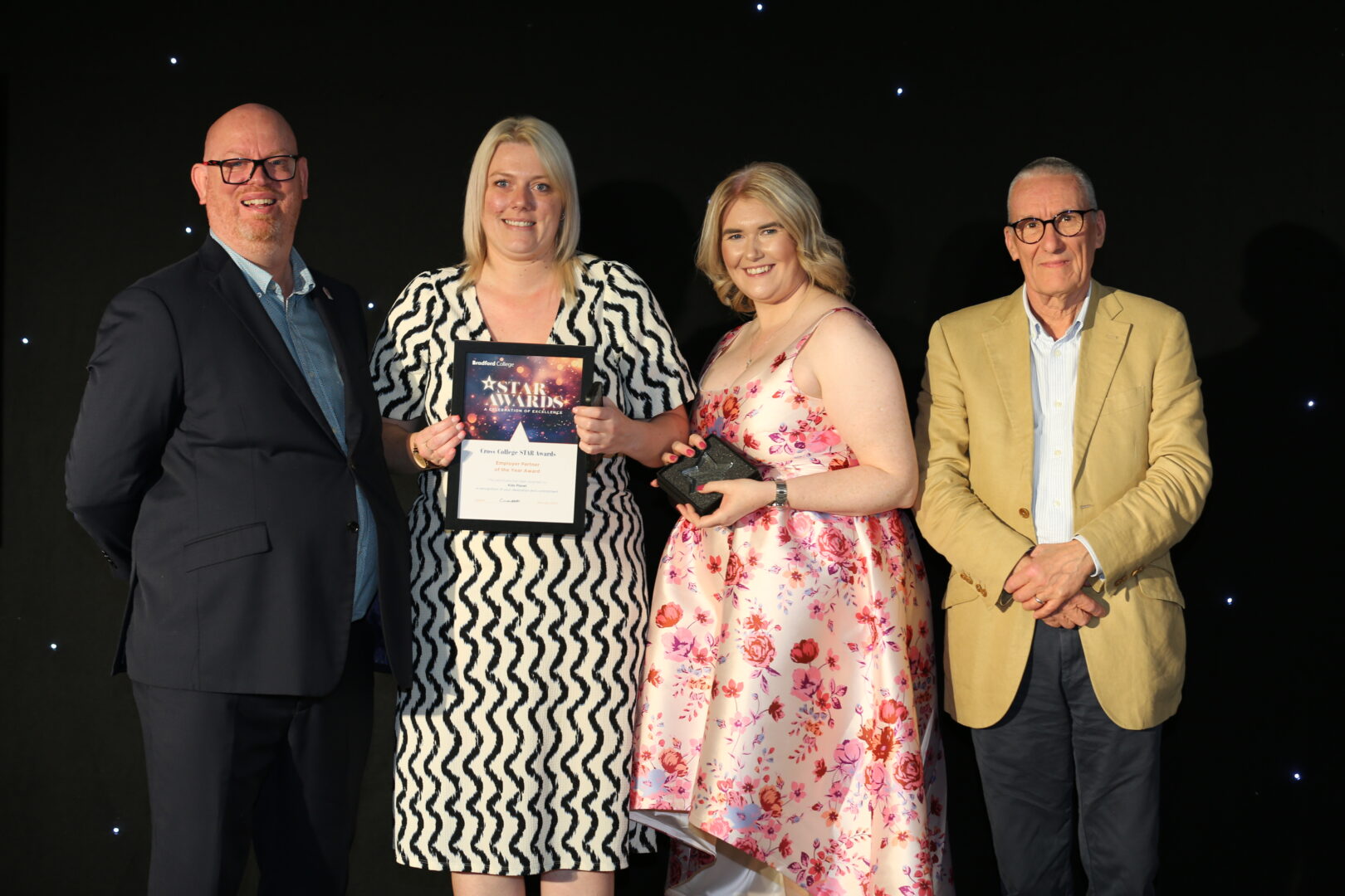 two award winners stand on stage holding certificates and awards