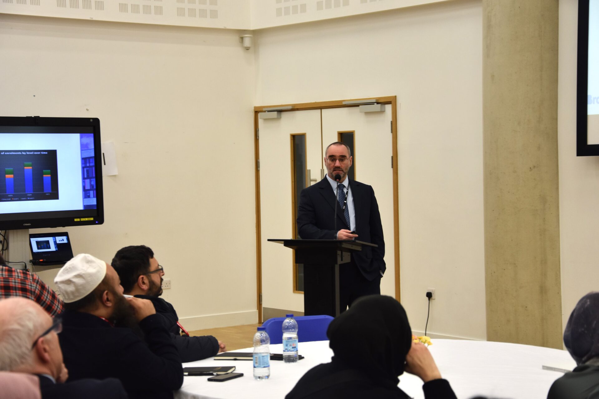 A male speaker stands at a podium in a conference room, presenting to an audience.