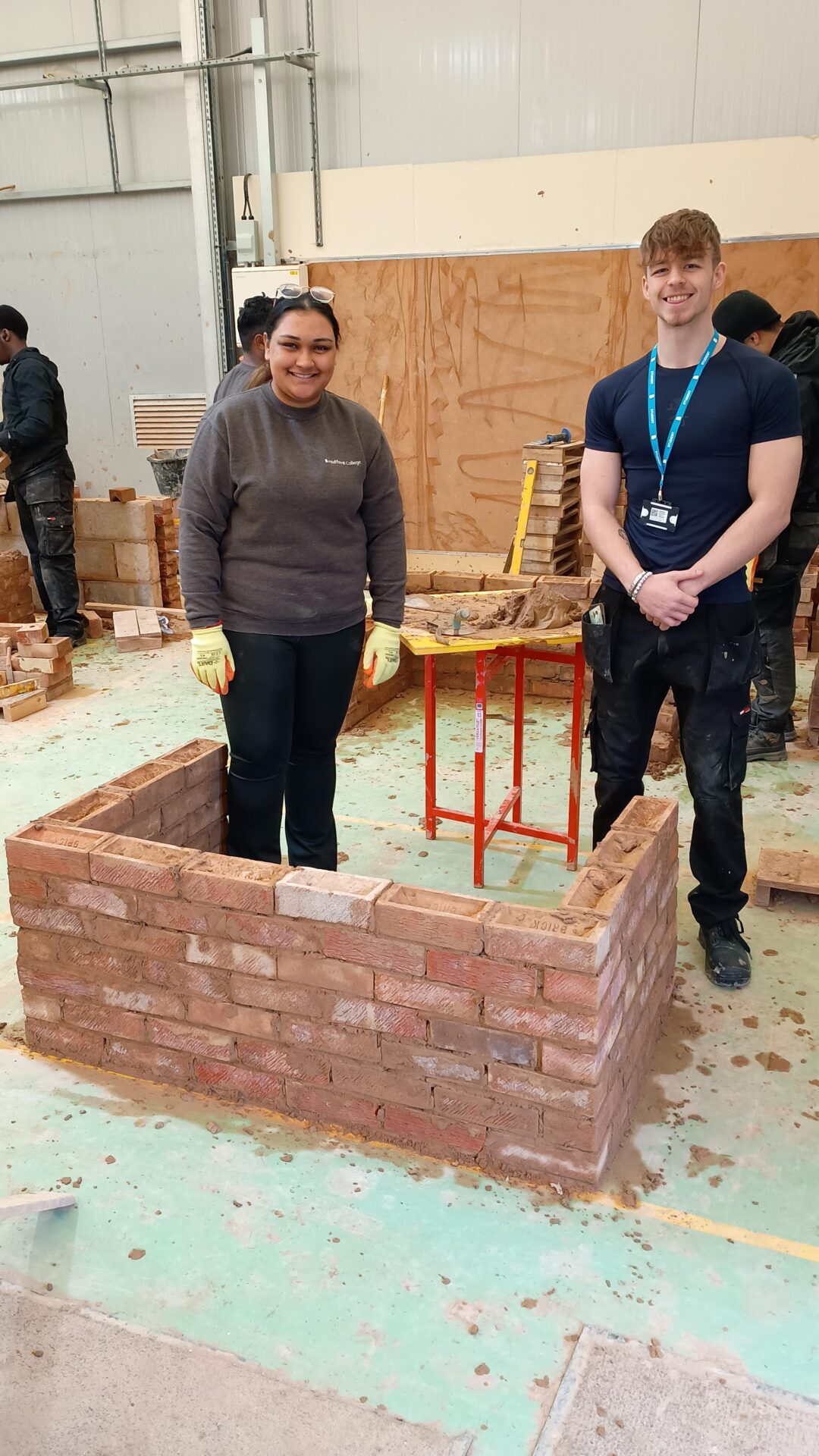 Image of a male and female students stood in front of a park built brick wall
