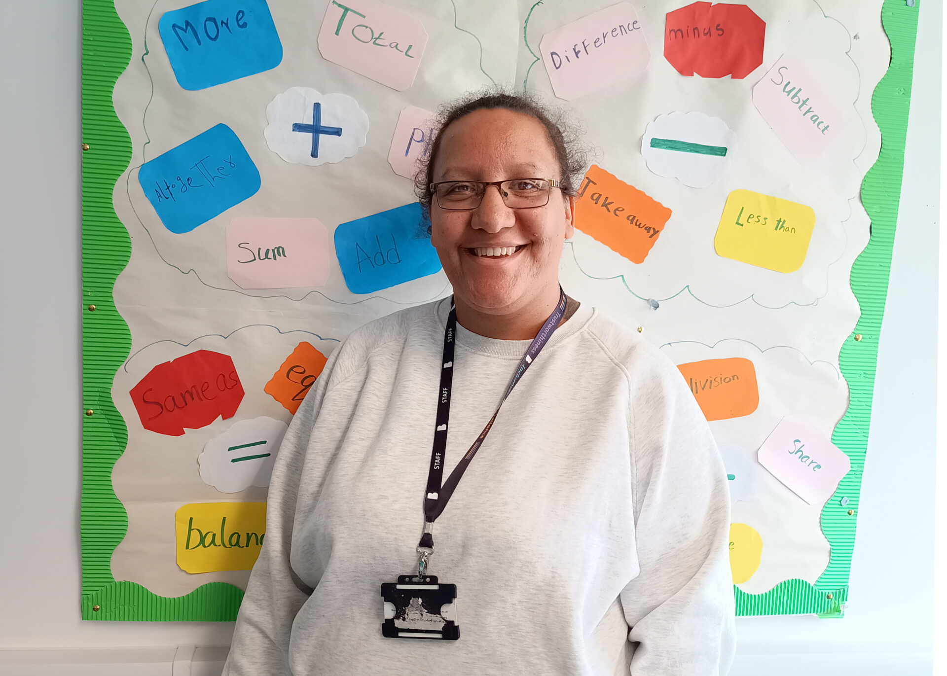 A person stands smiling in front of a colorful classroom-style display board decorated with cut‑out shapes containing handwritten words.