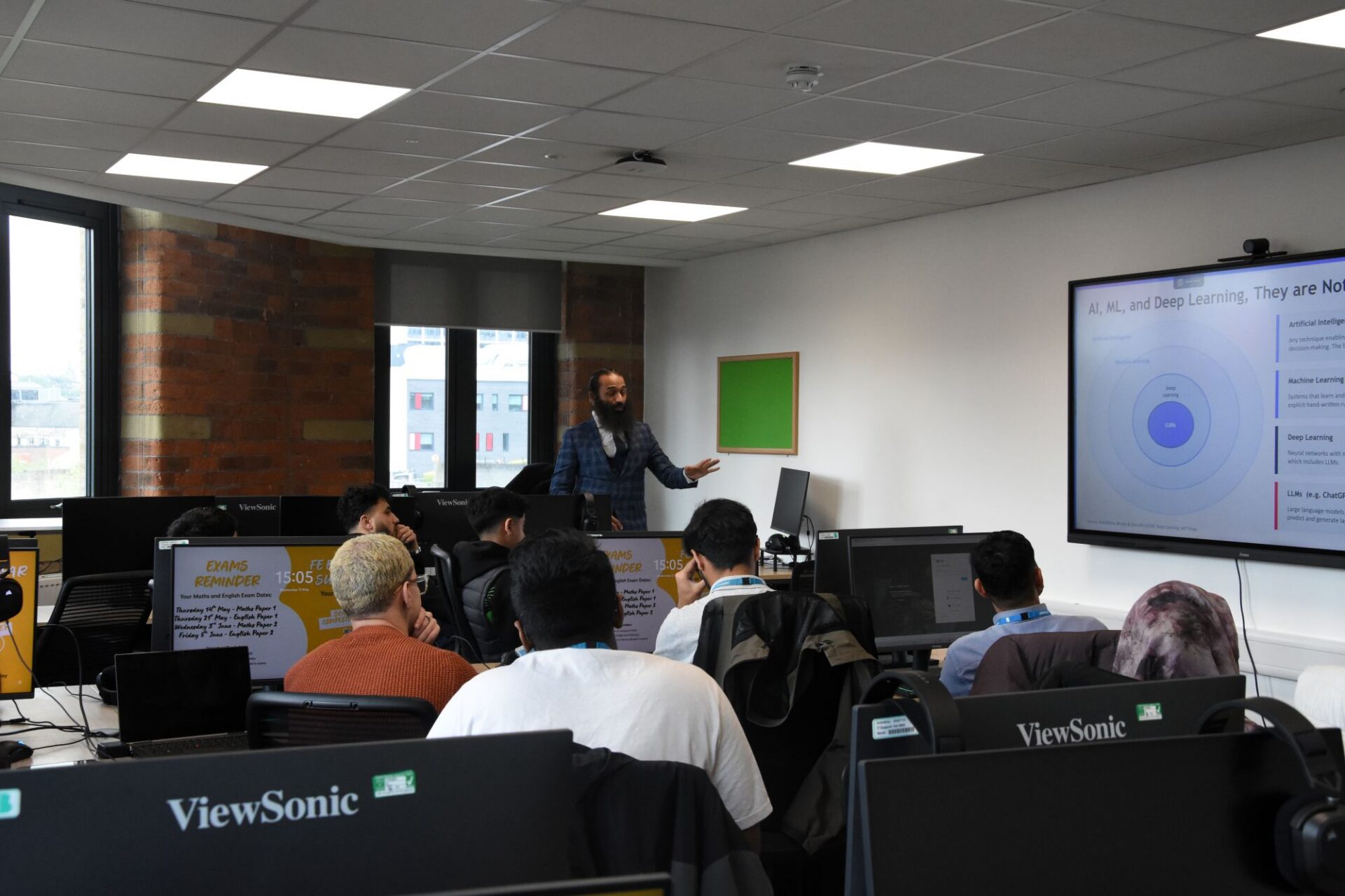A man stands in front of a computer talking to a class of students sat at desks listening.