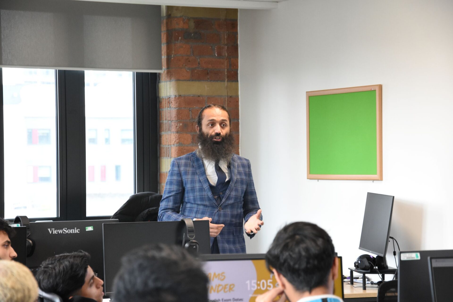 A man stands in front of a computer talking to a class of students sat at desks listening.