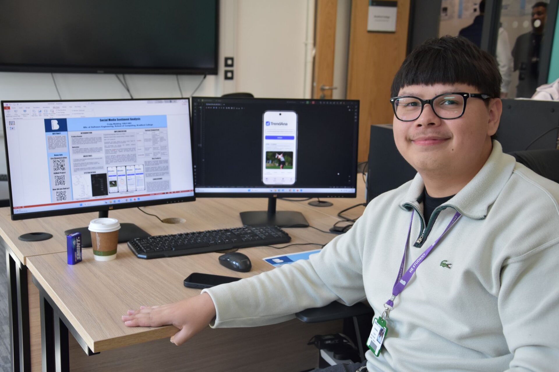 A male student sits at his desk in a classroom in front of his end of year project on his computer screen, smiling.