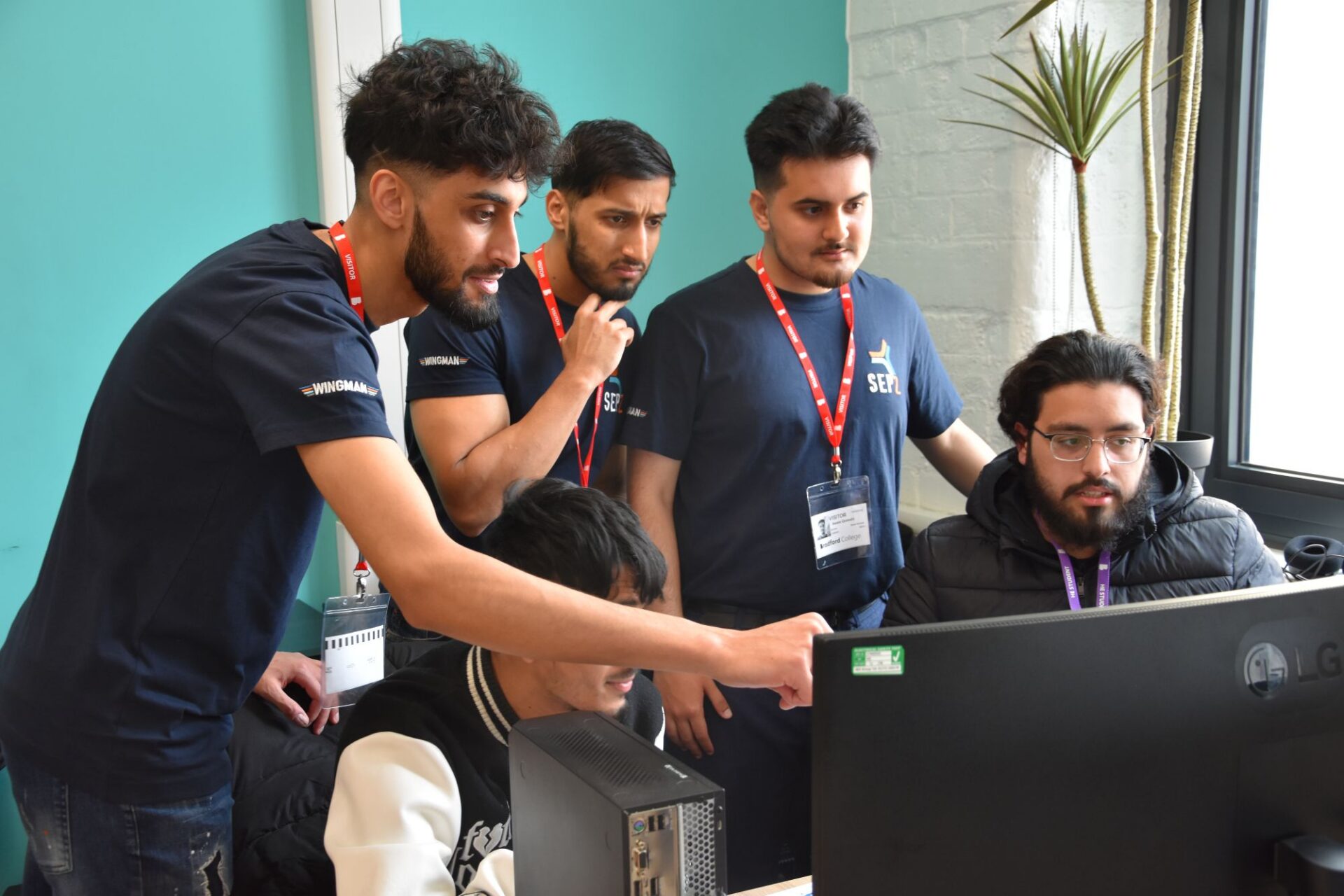 One man sits in front of his computer screen while 4 men stand beside him looking at the screen. One is pointing at the screen and talking.