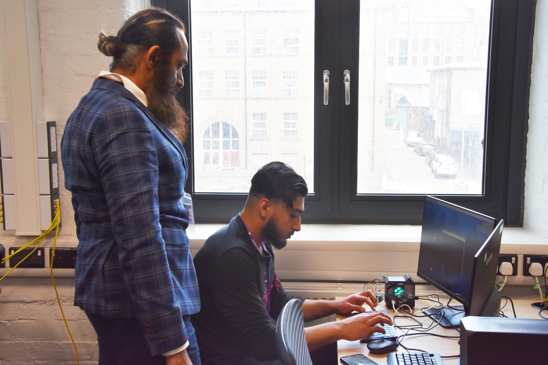 A male student works at his computer in a classroom while a man stands beside him looking at his screen.