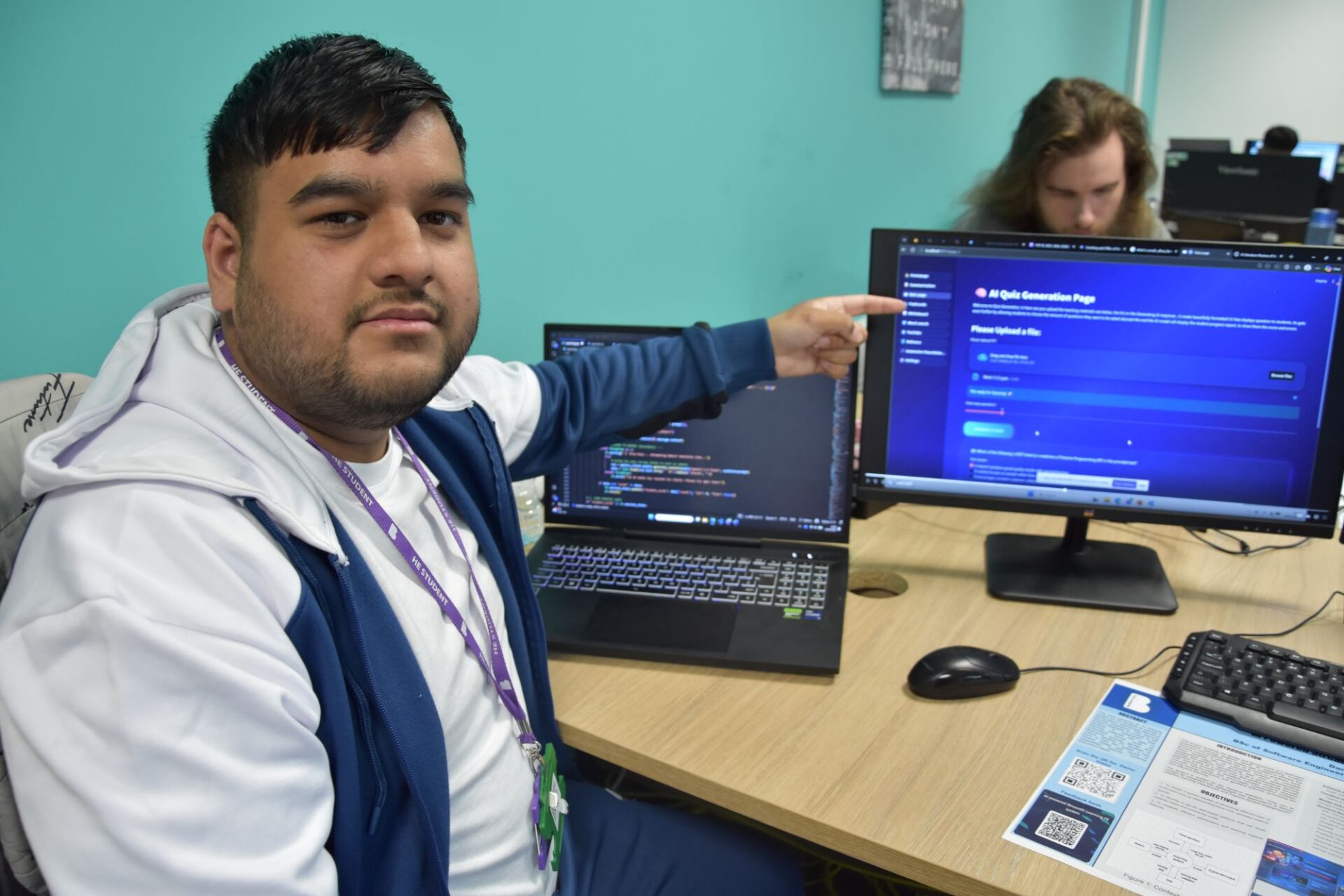A male student sits at his desk in a classroom pointing at a feature of his work on his computer screen.