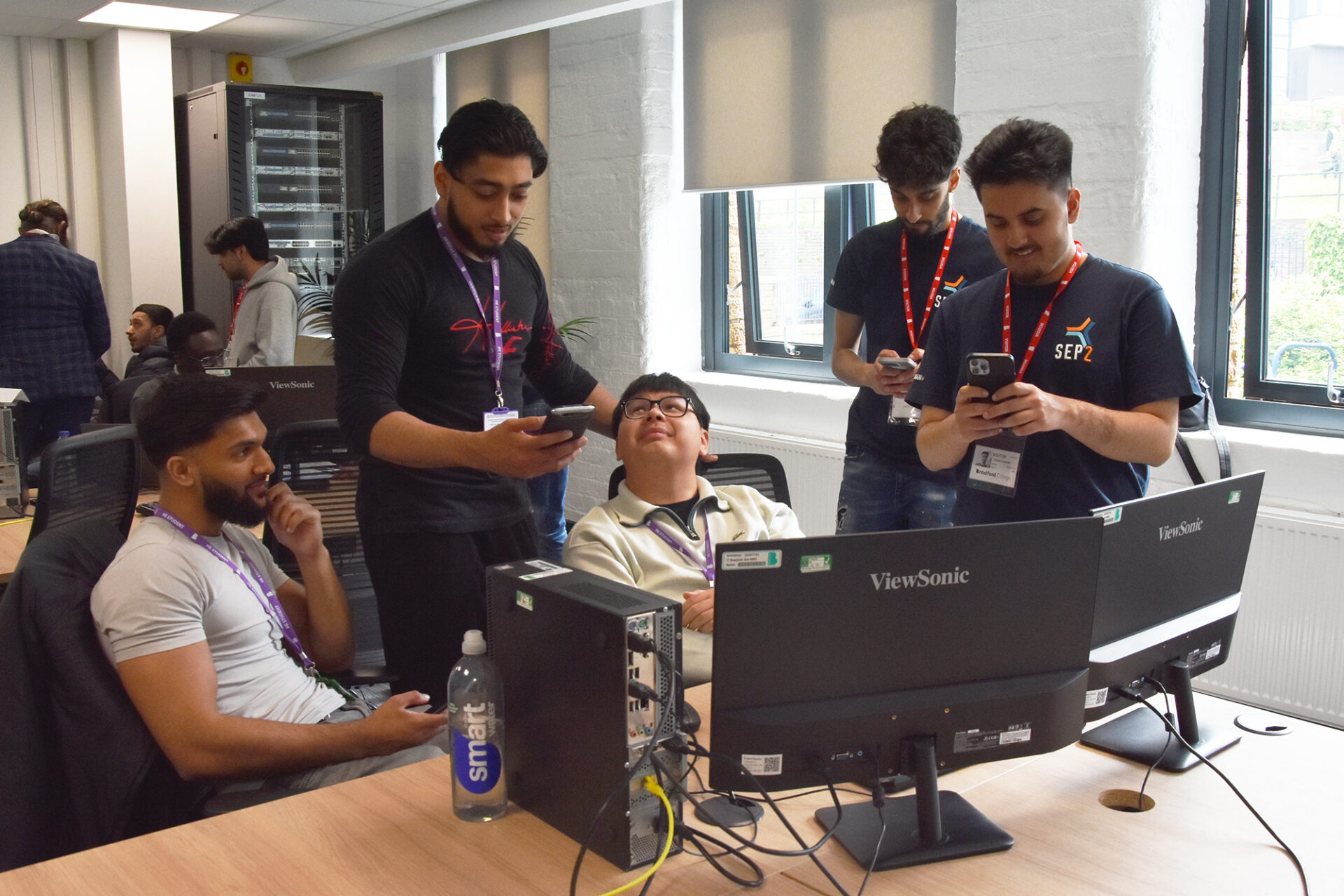 A group of men sit looking at a computer screen and their phone screens in a classroom.