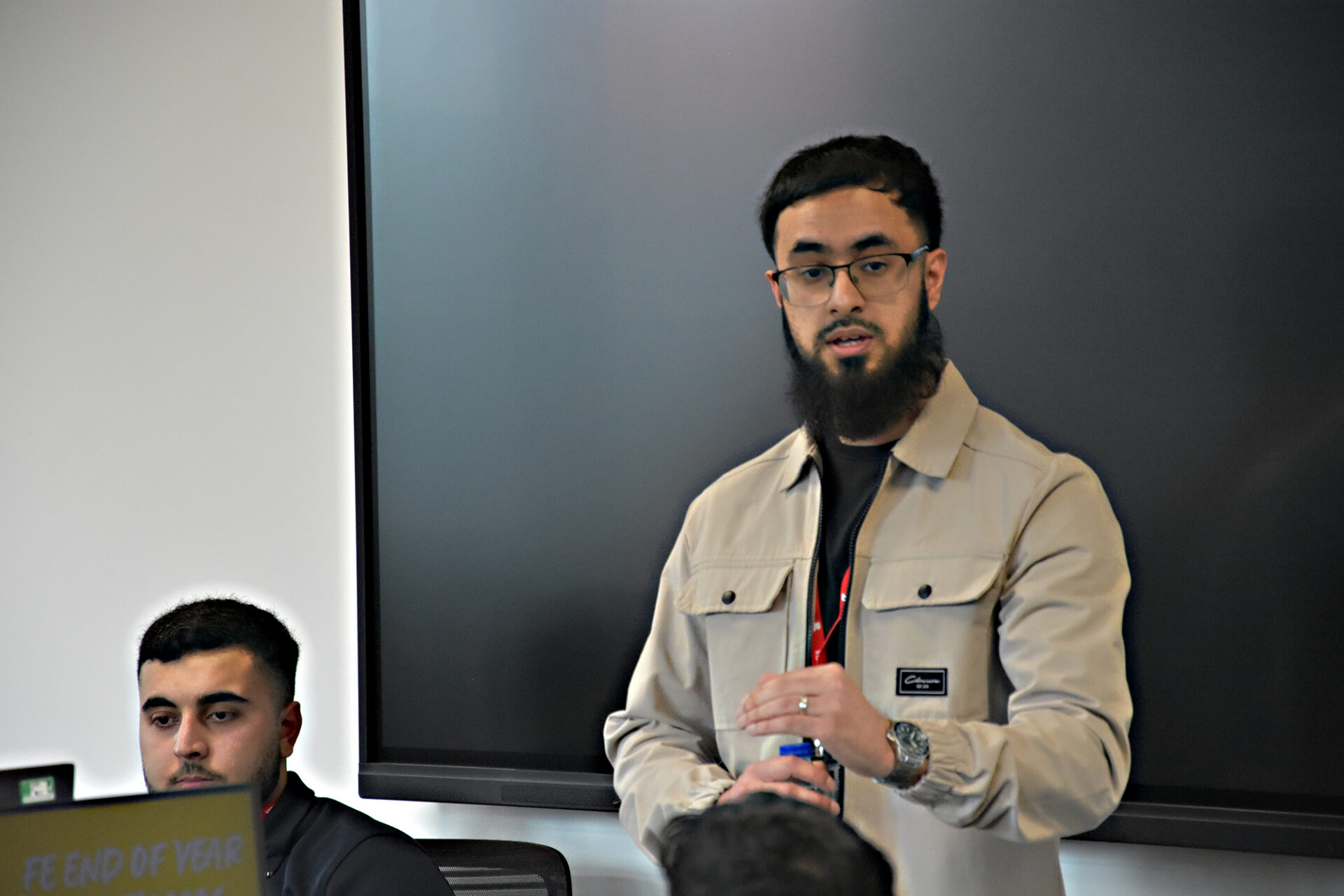 A man stands talking at the front of a classroom while students sit at desks with computers listening.