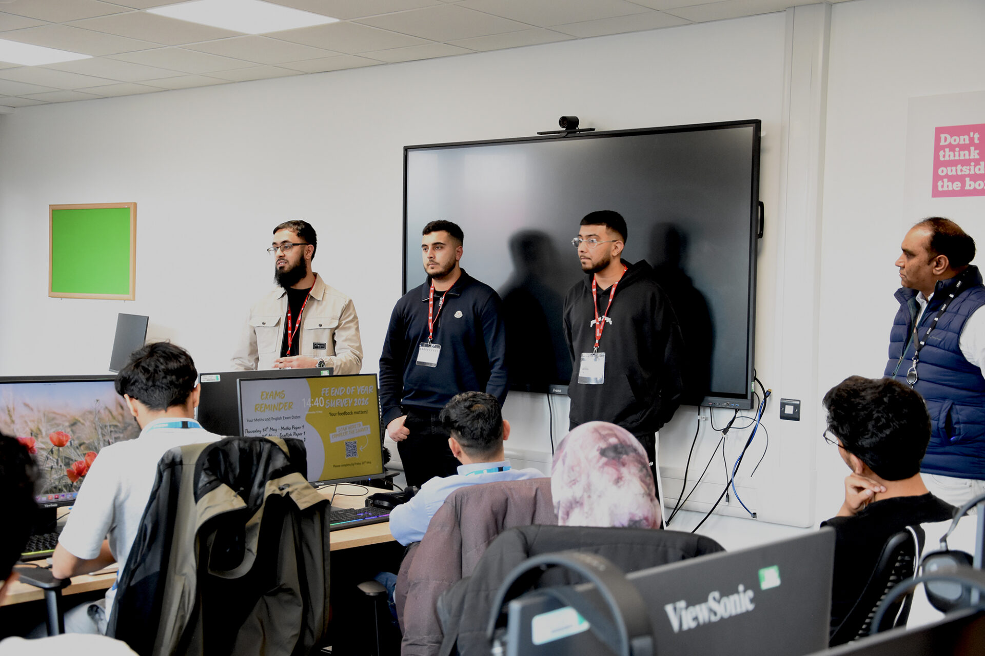 Four men stand talking at the front of a classroom while students sit at desks with computers listening.