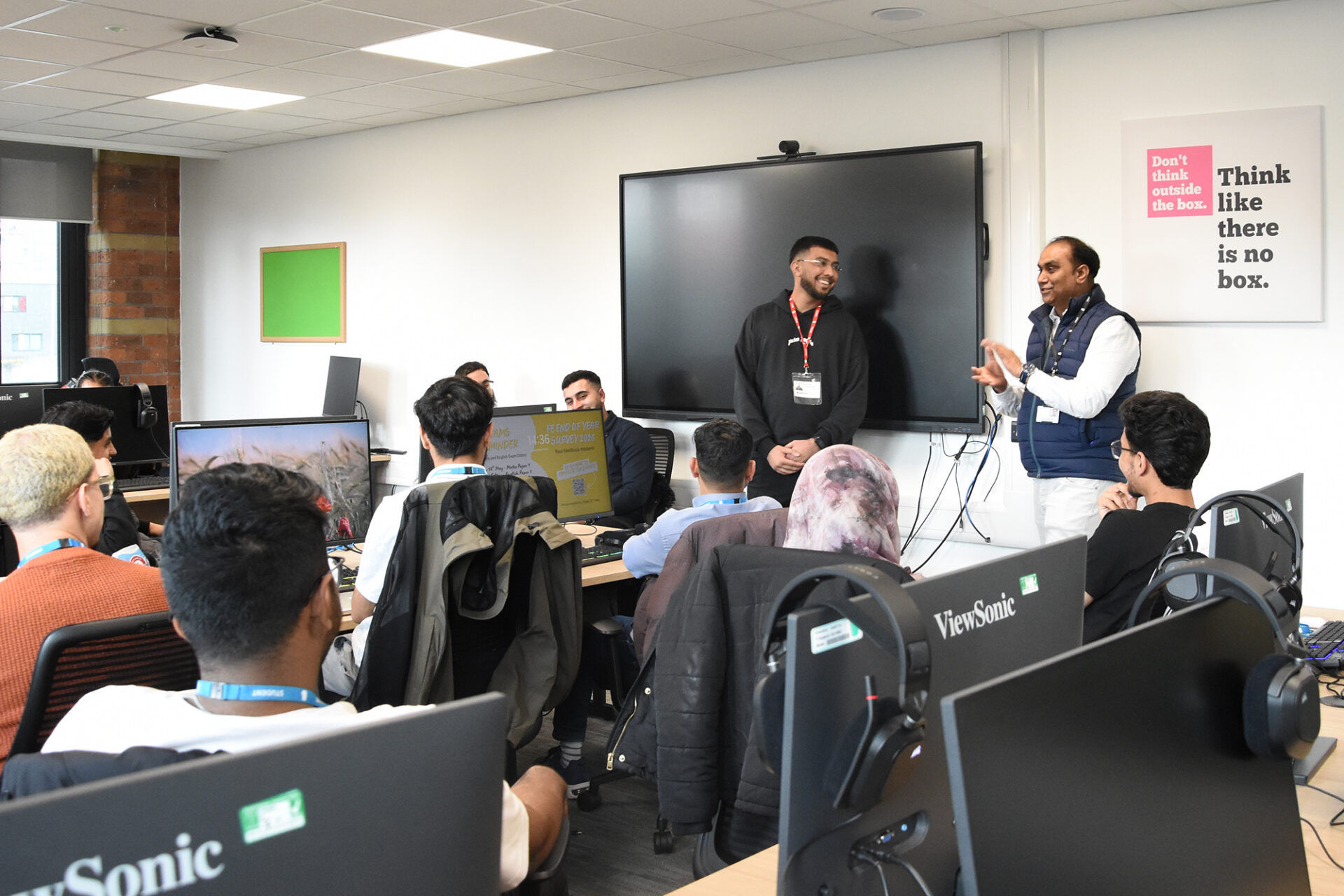 Two men stand talking at the front of a classroom while students sit at desks with computers listening.