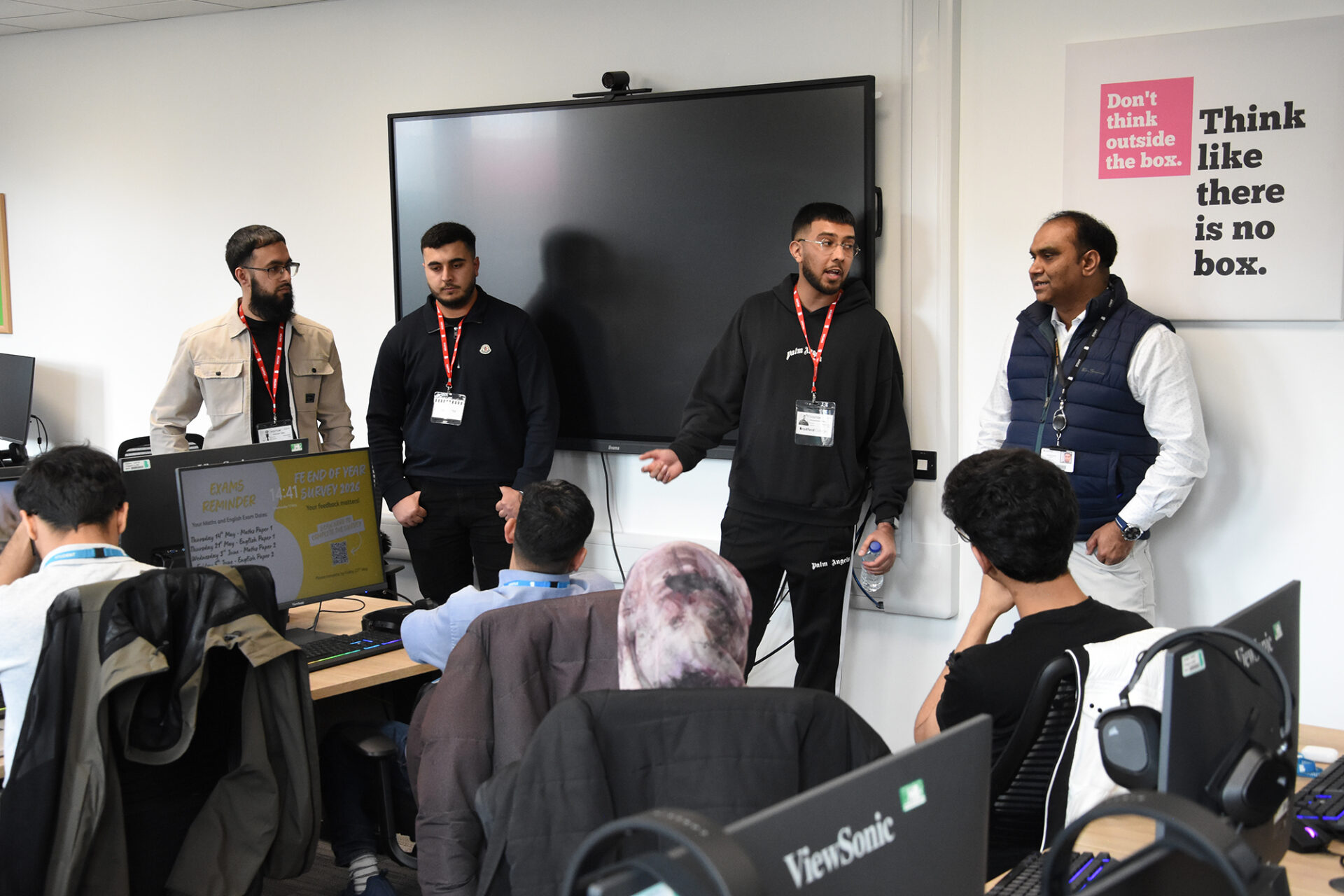 Four men stand talking at the front of a classroom while students sit at desks with computers listening.