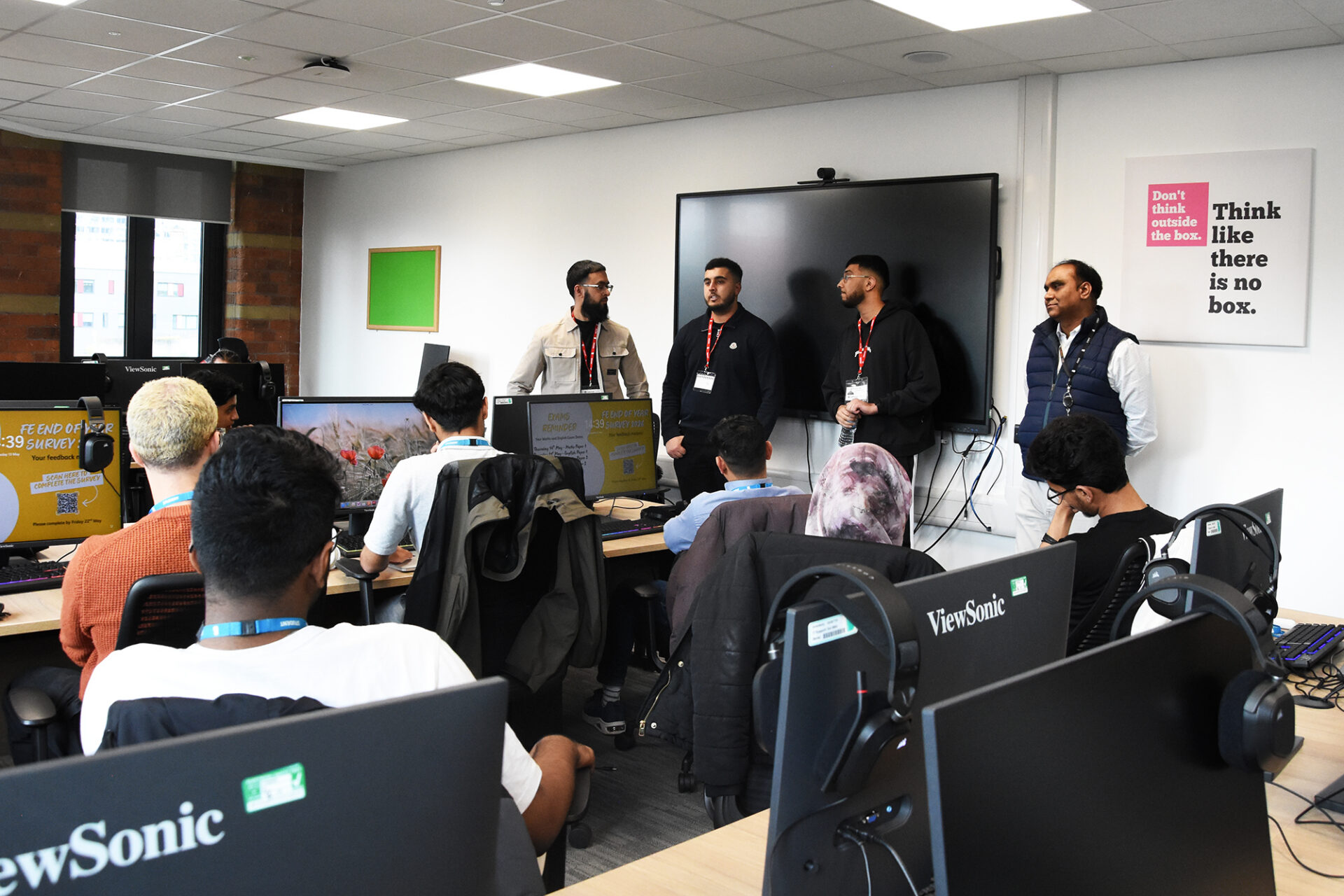 Four men stand talking at the front of a classroom while students sit at desks with computers listening.
