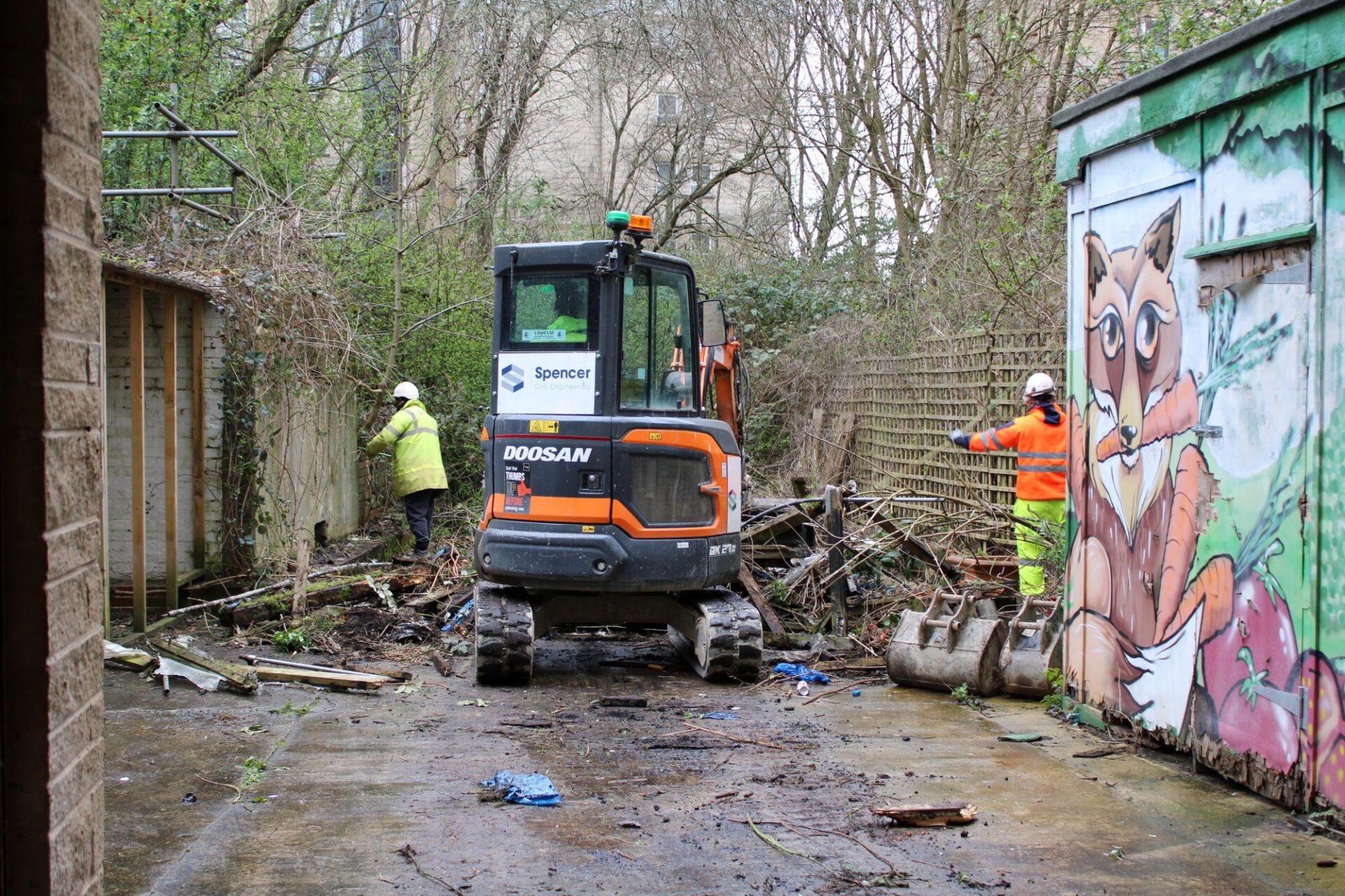 2 men wearing high vis jackets and hard hats, gloves and safety glasses work alongside a mini digger clearing an outdoor space.