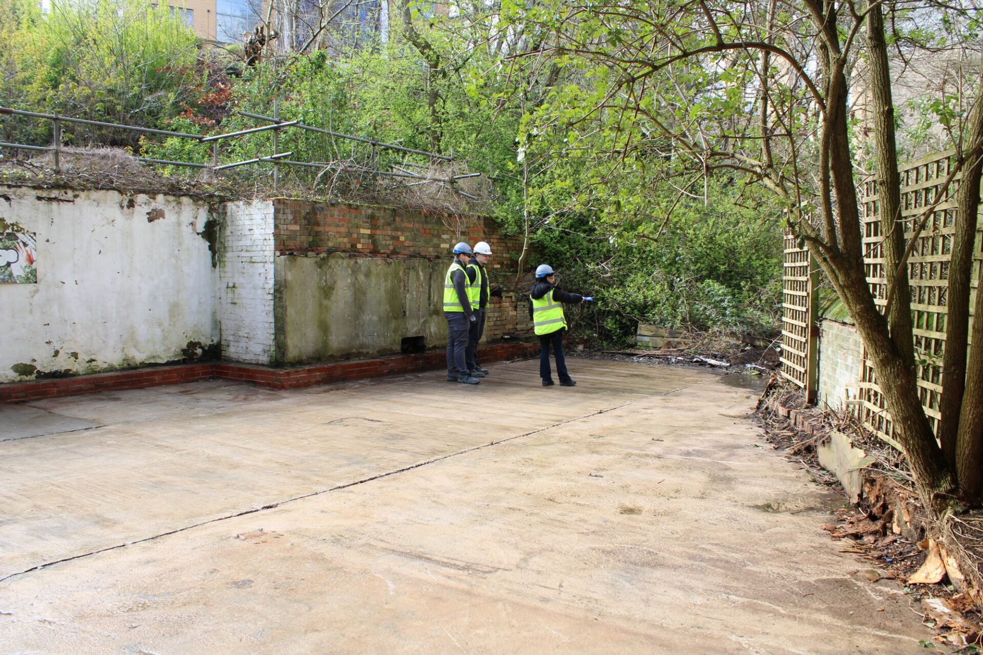A group of 2 men and 1 woman wearing high vis jackets and hard hats stand in a clear outdoor space pointing and talking.