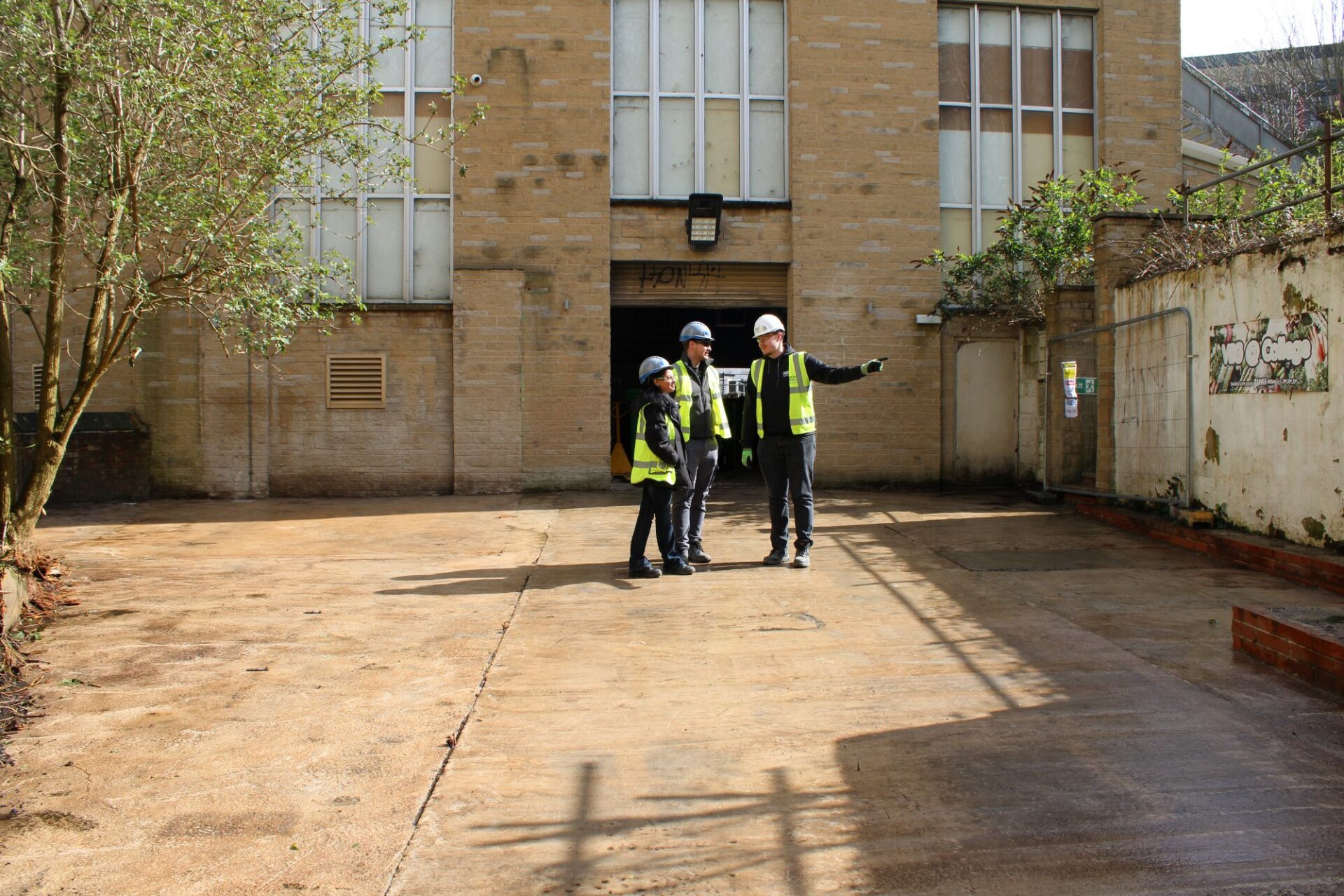 A group of 2 men and 1 woman wearing high vis jackets and hard hats stand in a clear outdoor space pointing and talking.