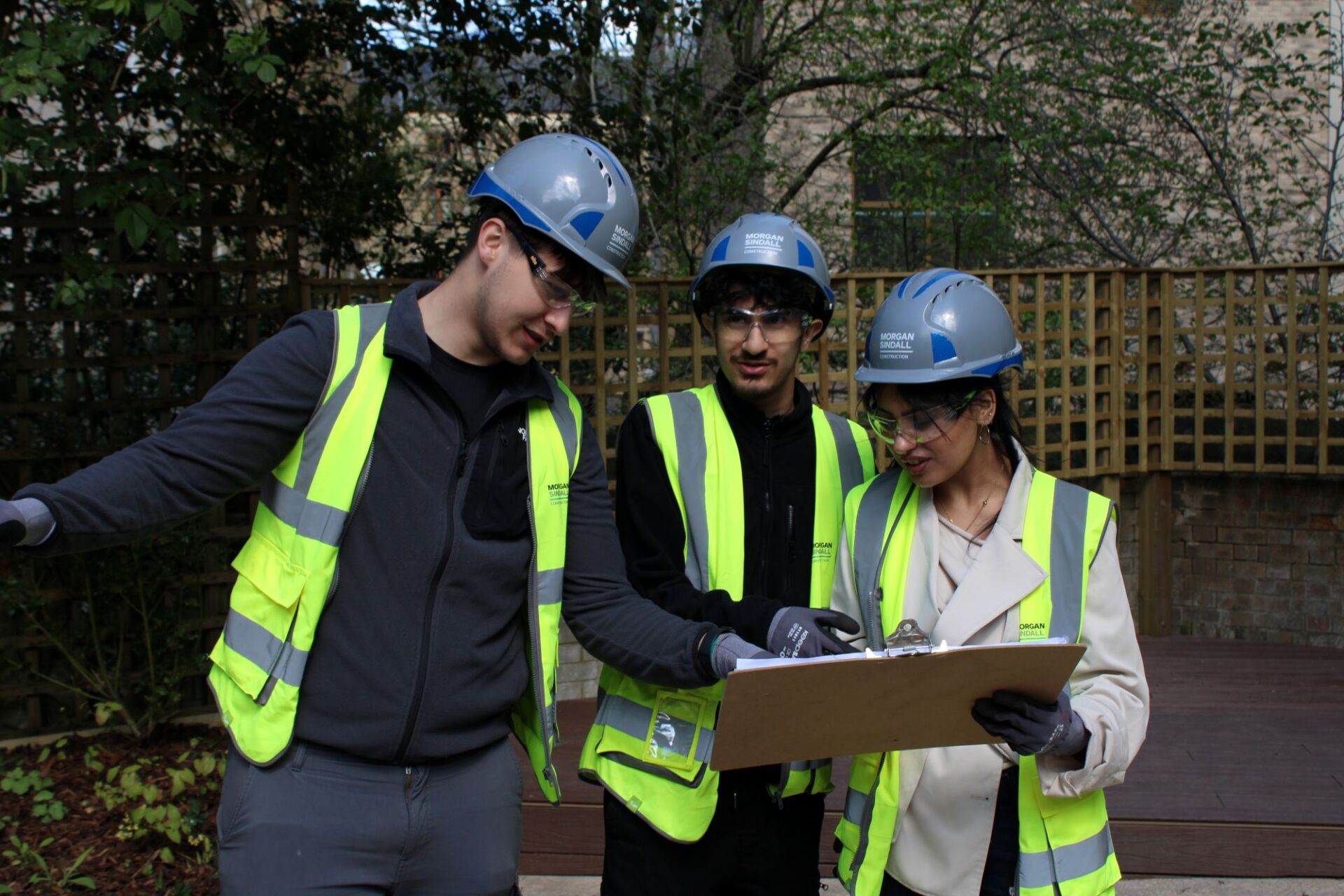 A group of 2 men and 1 woman wearing high vis jackets and hard hats, gloves and safety glasses stand together looking at a clipboard and pointing.