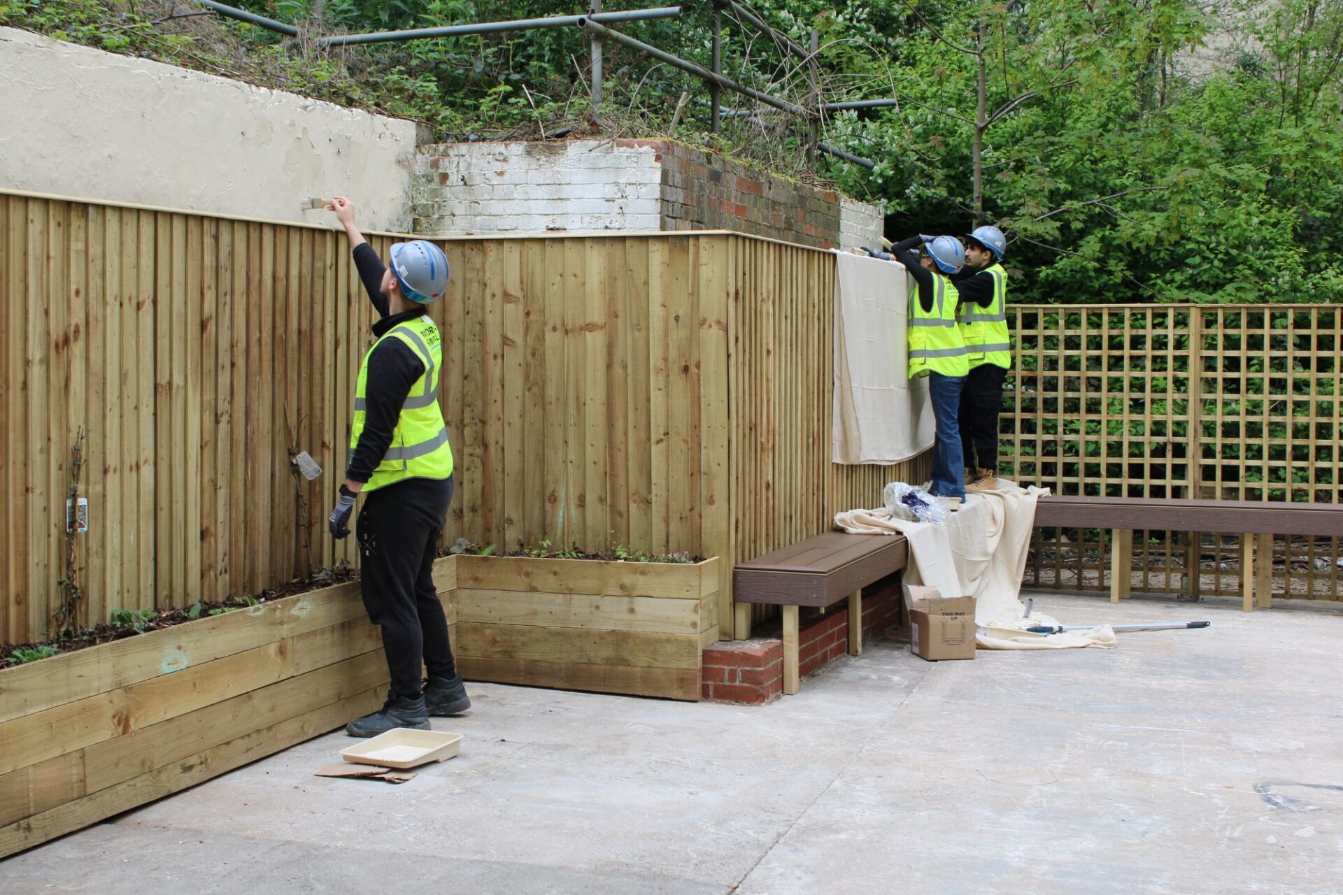 A group of 2 men and 1 woman wearing high vis jackets and hard hats paint a garden wall cream.