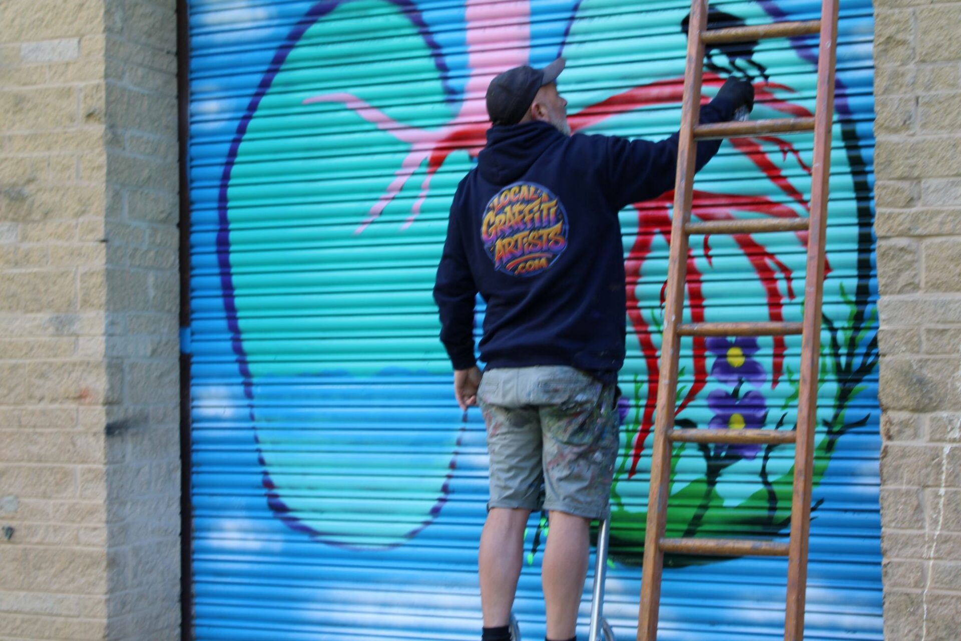 A man stands on a step ladder spray painting a mural of flowers and nature filling some lungs, on a roller shutter door.