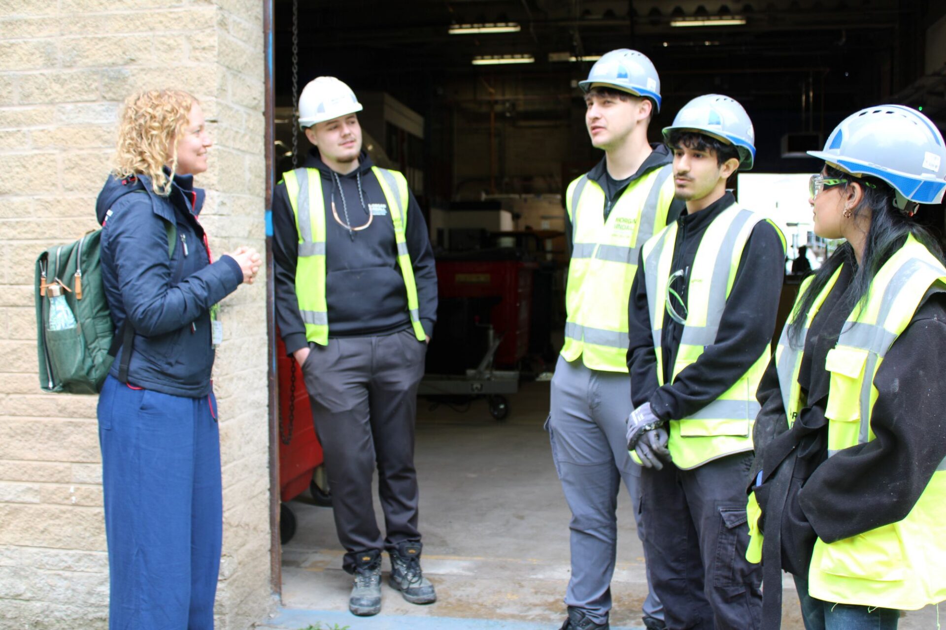 A group of 3 men and 1 woman wearing high vis jackets and hard hats stand together talking to a visitor about the garden design.