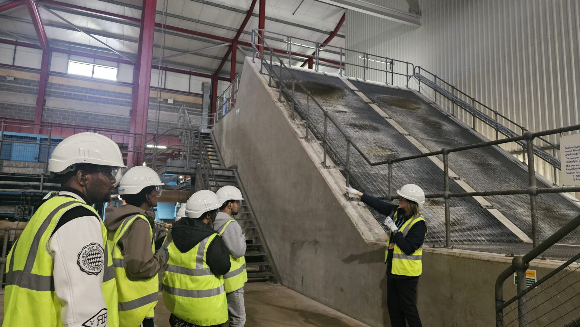 A group of 3 men and 2 women stand listening to a woman showing them a facility at Yorkshire Water. The group are all wearing high vis jackets, gloves, safety glasses and hard hats.