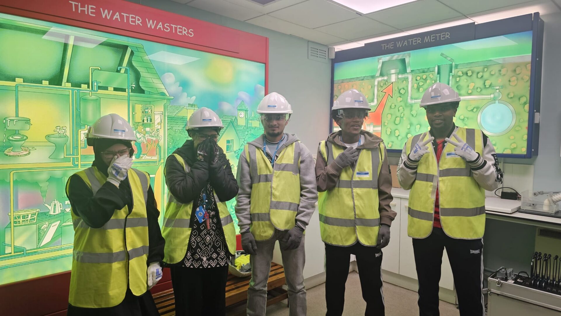 A group of 3 men and 2 women stand in front of large illuminated signs showing illustrations entitle 'the water meter' and 'the water wasters'. They are all wearing high vis jackets, gloves, safety glasses and hard hats.