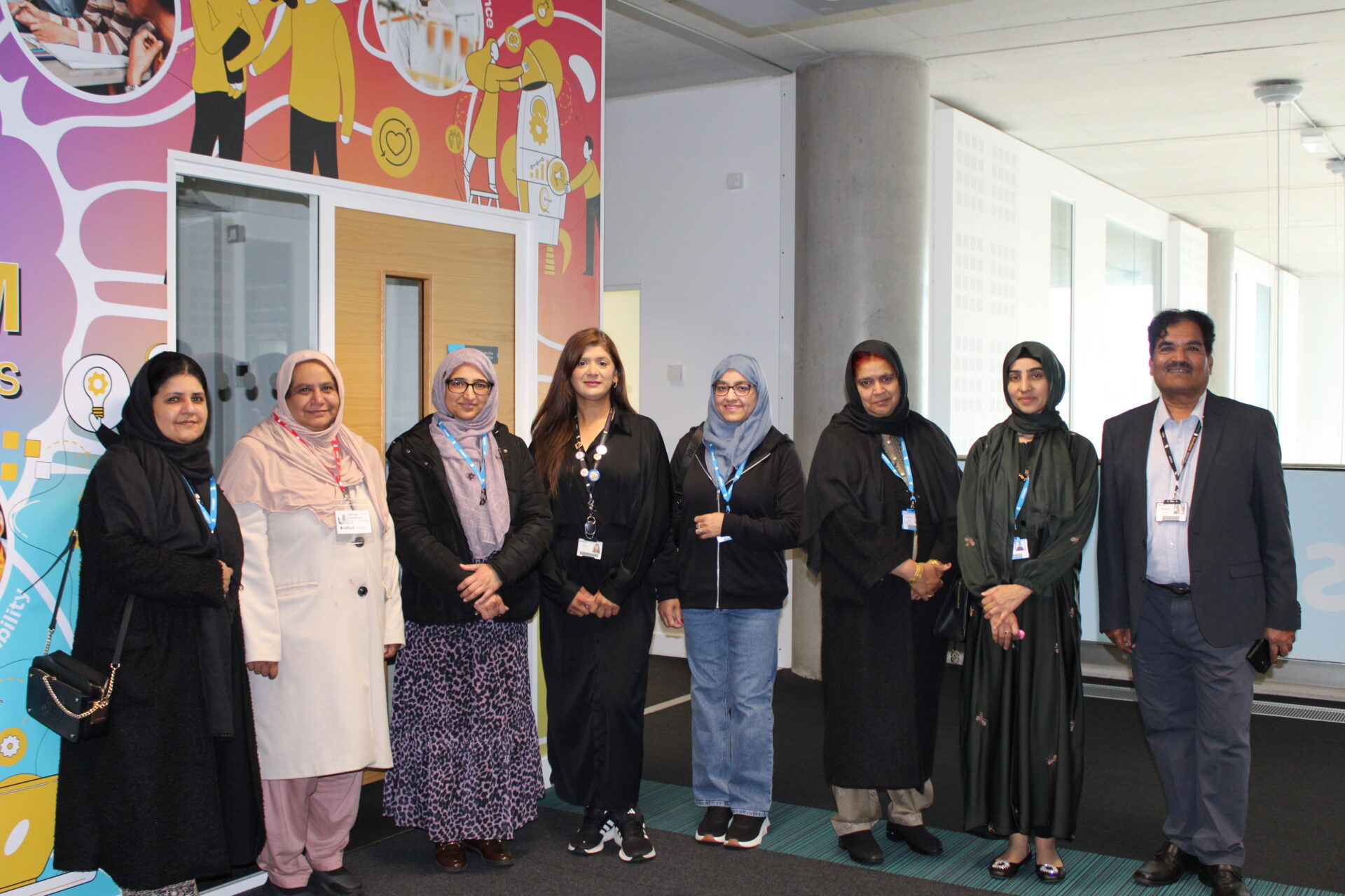 A group of nine adults stand in a line inside a modern college building, facing the camera. Several wear lanyards, and some wear head coverings. A colourful illustrated wall is on the left, with a doorway and large windowed corridor extending to the right.