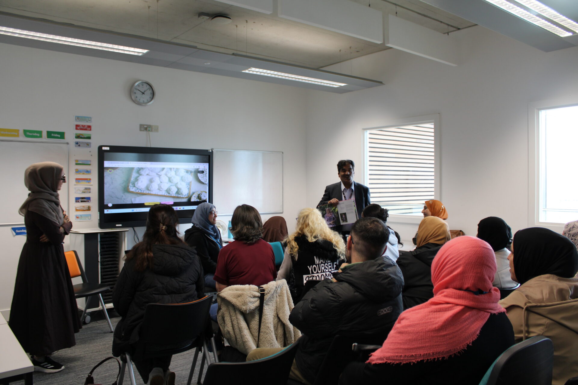 A classroom session in progress, with a group of adult learners seated and facing a speaker at the front. The speaker holds up an open book while standing beside a large screen displaying food images. A whiteboard, wall clock, and labelled classroom displays are visible.