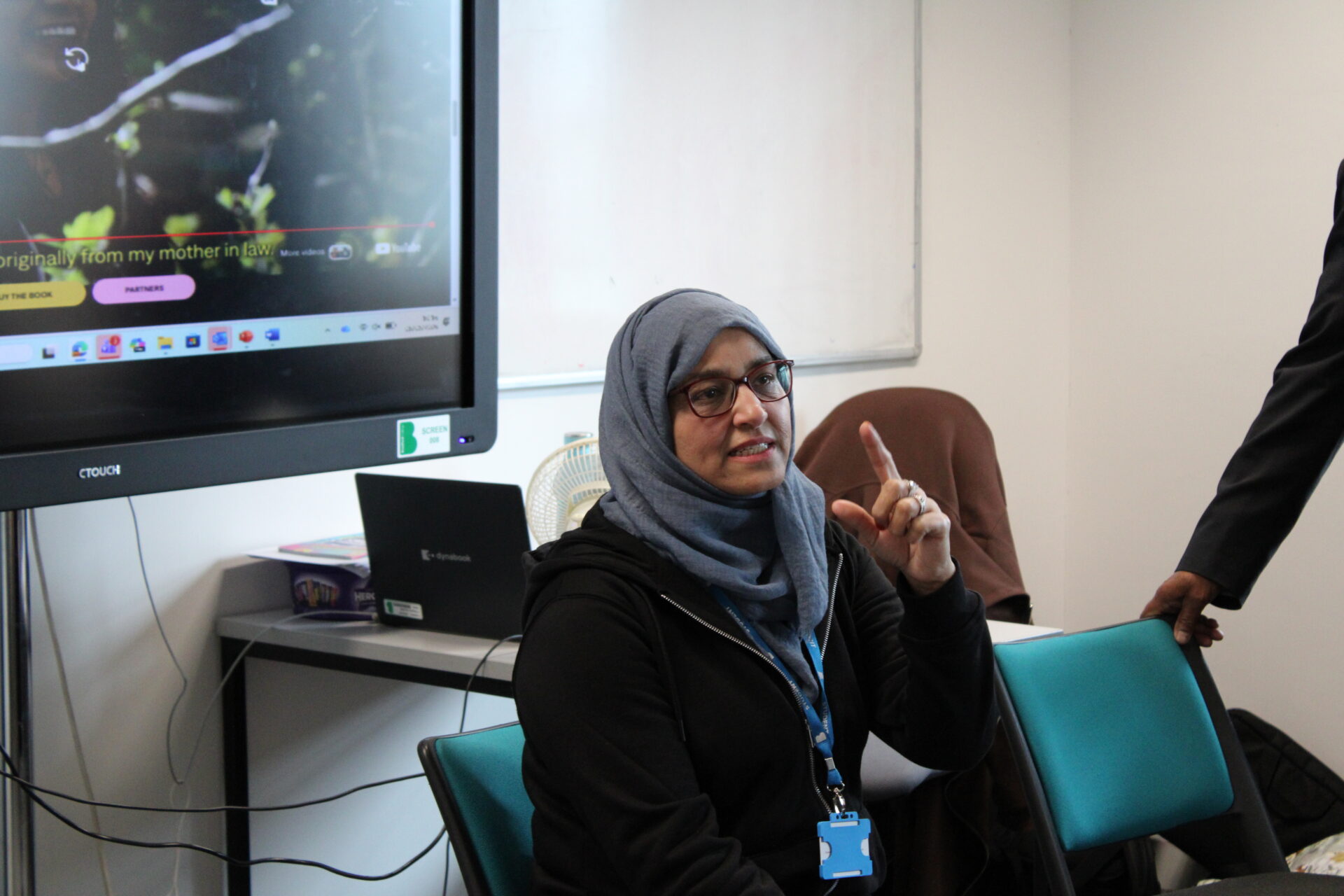 An adult learner sits in a classroom, raising a finger while speaking during a discussion. A large interactive screen showing a webpage is visible beside them, along with a laptop, whiteboard, and classroom equipment in the background.  