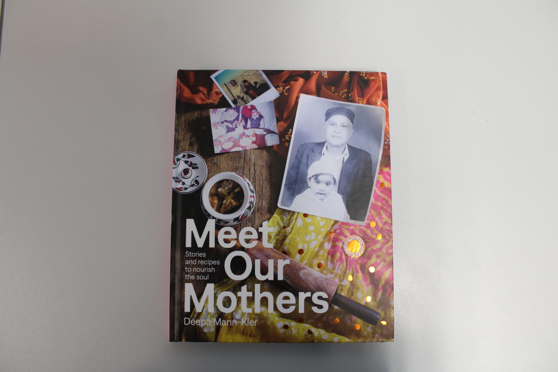 A copy of the book Meet Our Mothers lies on a white surface. The cover features a collage of photographs, including family images, a black and white portrait, and food items, alongside colourful fabric and the title text "Meet Our Mothers: Stories and recipes to nourish the soul" by Deepa Mann-Kler.