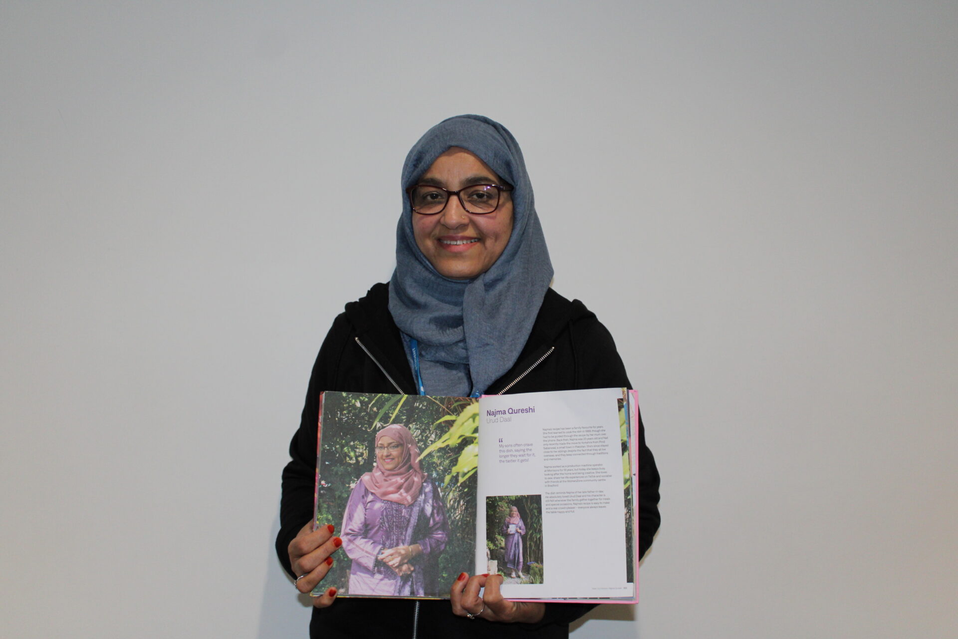 A person stands against a plain white background holding open a colourful cookbook. The page shows a large outdoor photograph and printed text, including the name “Najma Qureshi” and a recipe title. The person is wearing glasses, a head covering, and a lanyard, and is facing the camera.