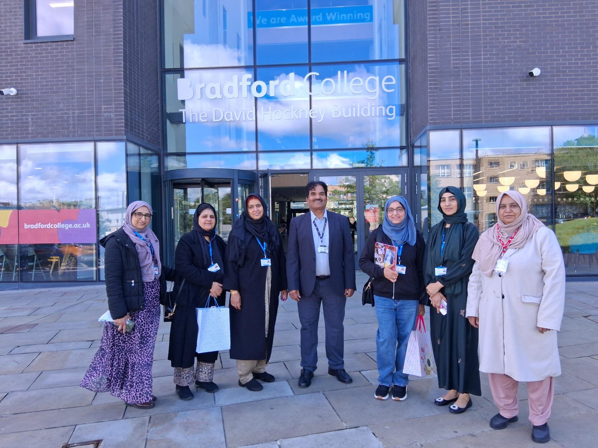 A group of eight adults stand outside the entrance of Bradford College's David Hockney Building, posing in front of large glass doors with the building name displayed above. Several individuals wear lanyards, and one person holds a colourful book. A Bradford College sign and reflections of the surrounding area are visible in the glass facade behind them. 