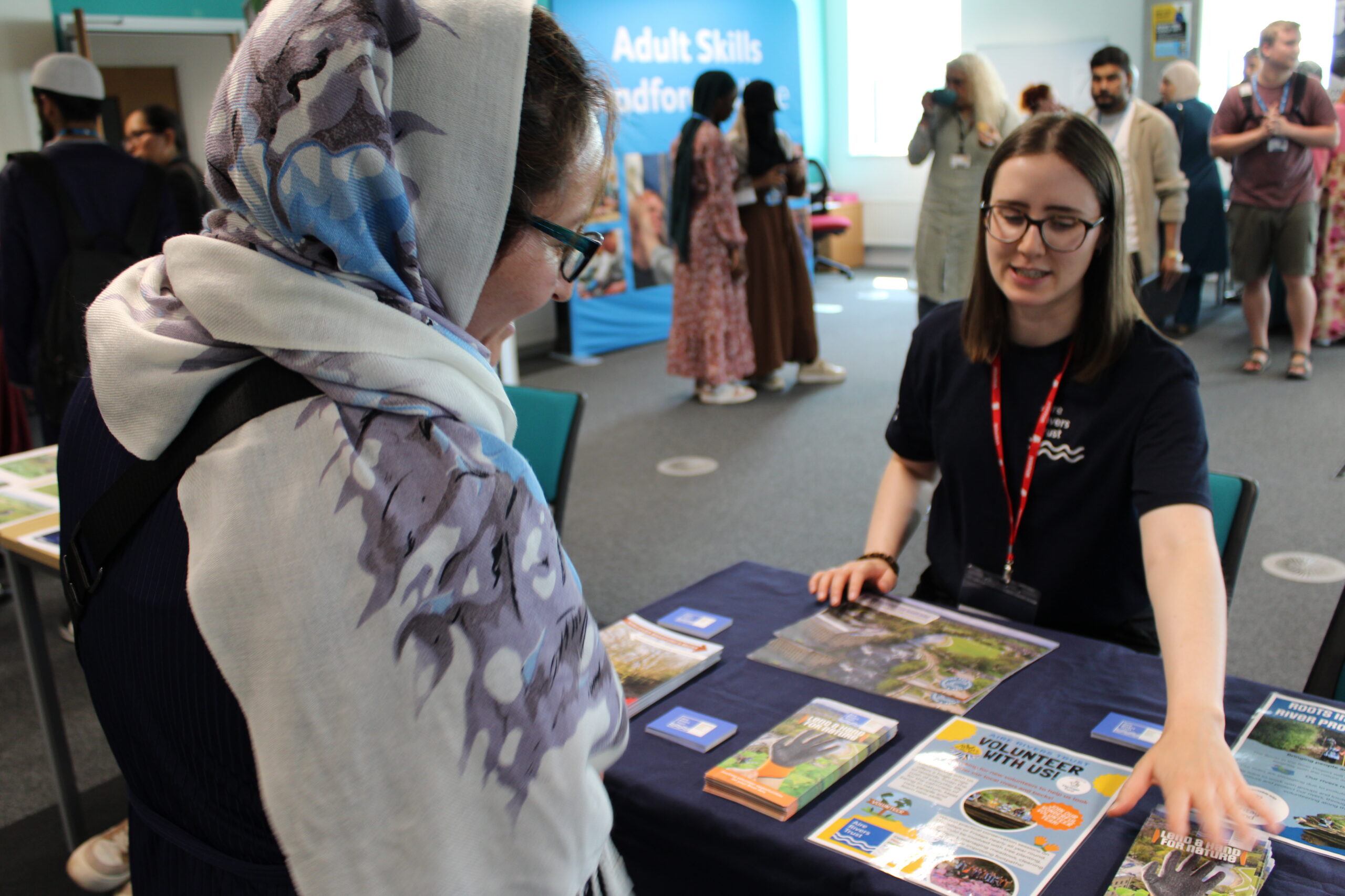 An adult learner and a visitor talk about volunteering while pointing at a leaflet. There are lots of other people at the event in the background.