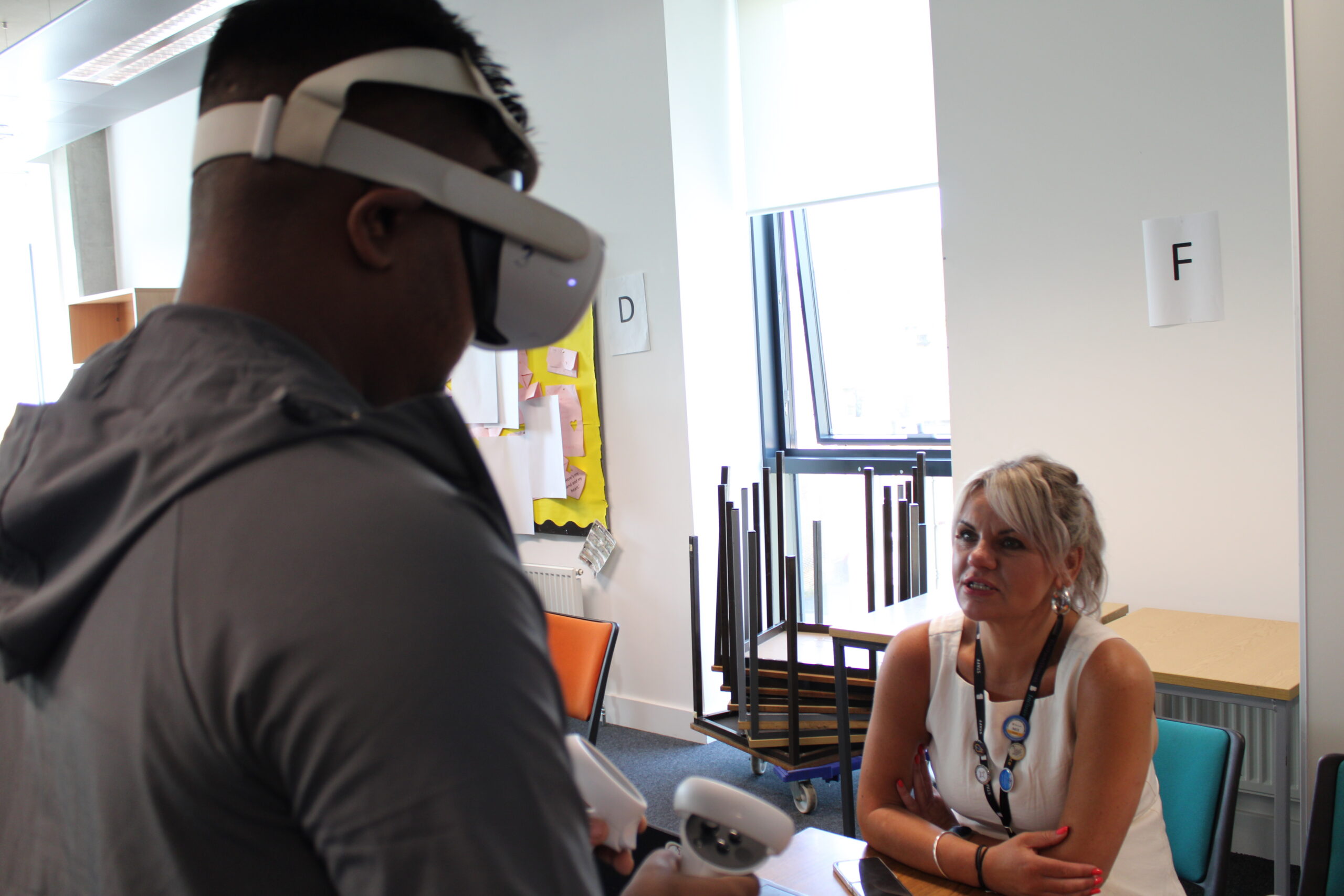 An adult learner speaks with a staff member while wearing a VR headset and holding the VR controllers in each hand.