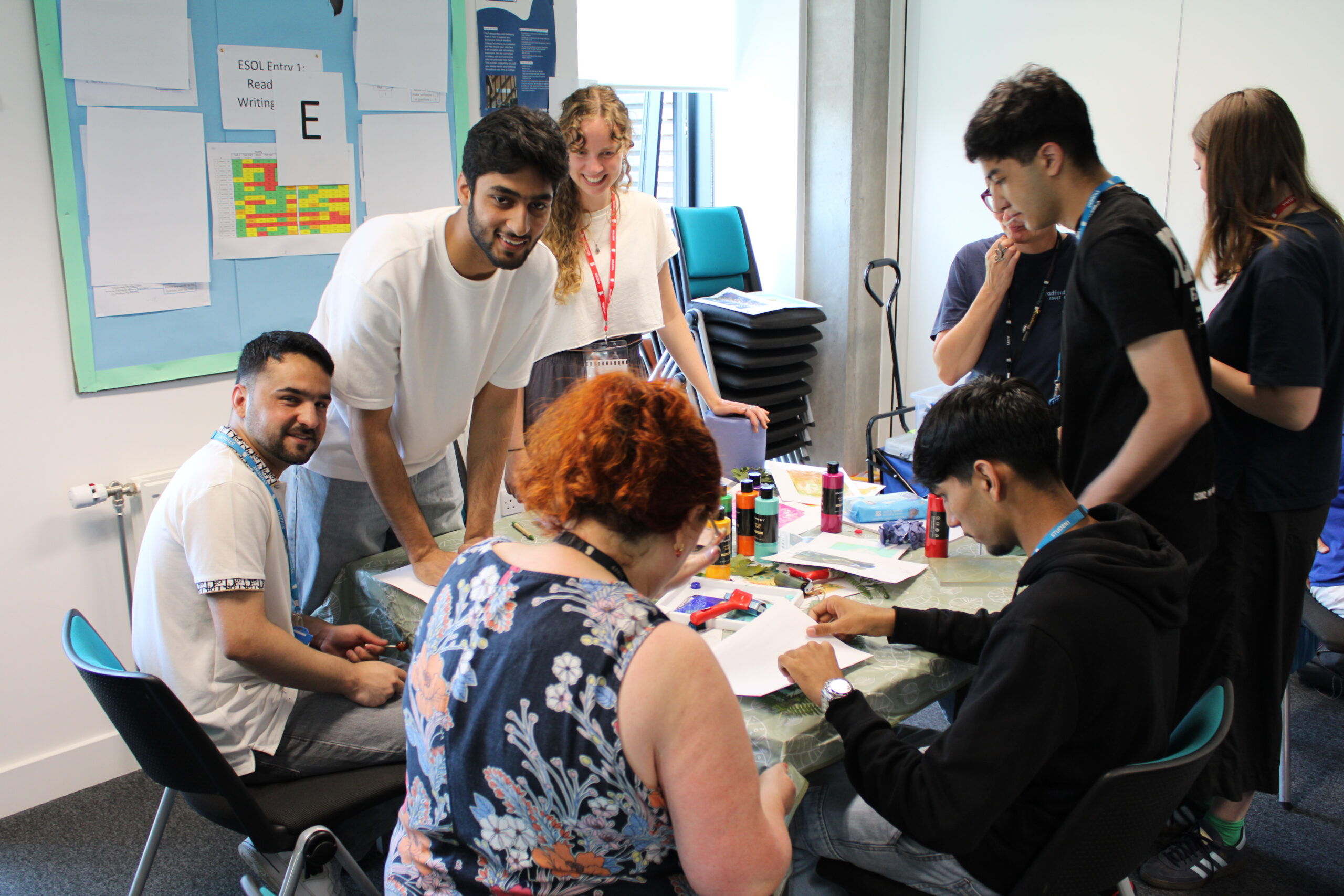 A group of adult students gather around a table covered with paints and materials to make leaf prints.