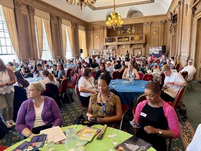 A large audience sits at round tables inside an ornate hall during an adult learning event. The room features wood-panelled walls, chandeliers, tall windows with curtains, and rows of red chairs. Attendees are seated throughout the space, with some reading event materials and others using mobile phones while listening to proceedings. The hall is busy and well attended, with a display board visible at the far end of the room.