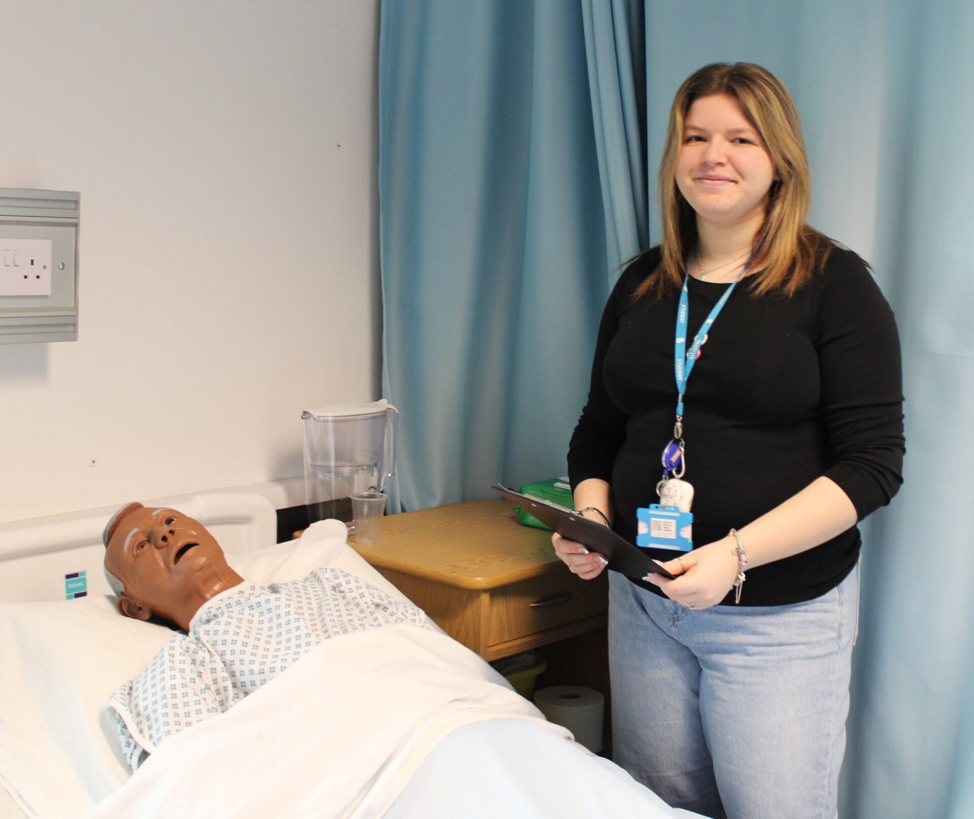 A person stands beside a hospital‑style bed holding a clipboard. In the background are light blue privacy curtains.